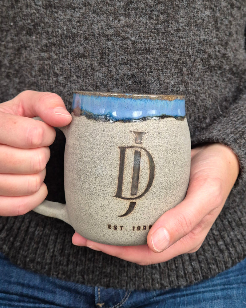 Person holding a ceramic mug with a logo and text, wearing a dark sweater.