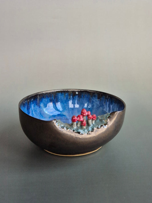 Small bowl featuring hand sculpted tiny toadstools  on the rim. Bronze glazed exterior, deep blue interior. On a grey background