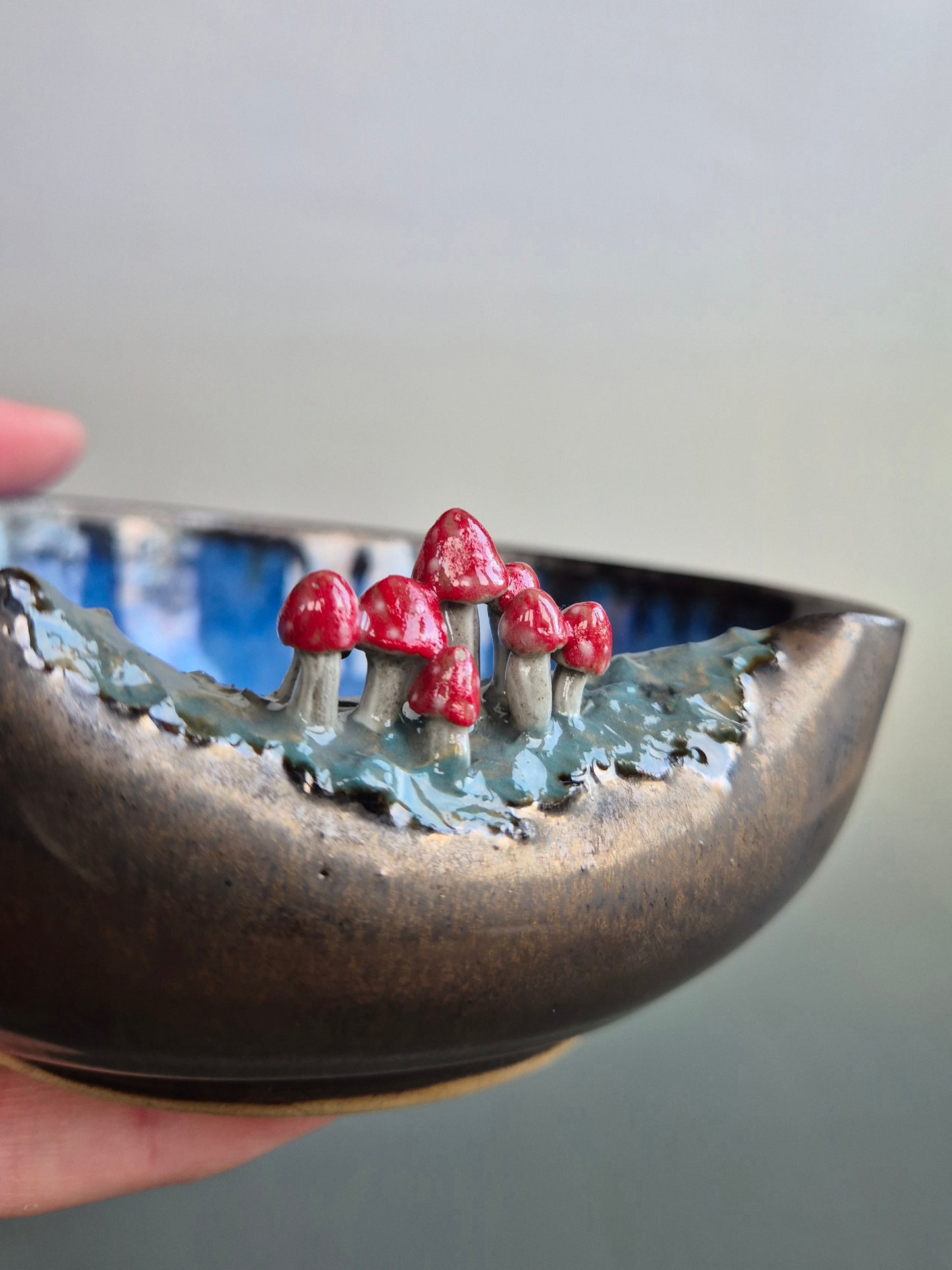 Close up of a small bowl featuring hand sculpted tiny toadstools  on the rim. Bronze glazed exterior. The bowl is resting in a person's hand. On a grey background