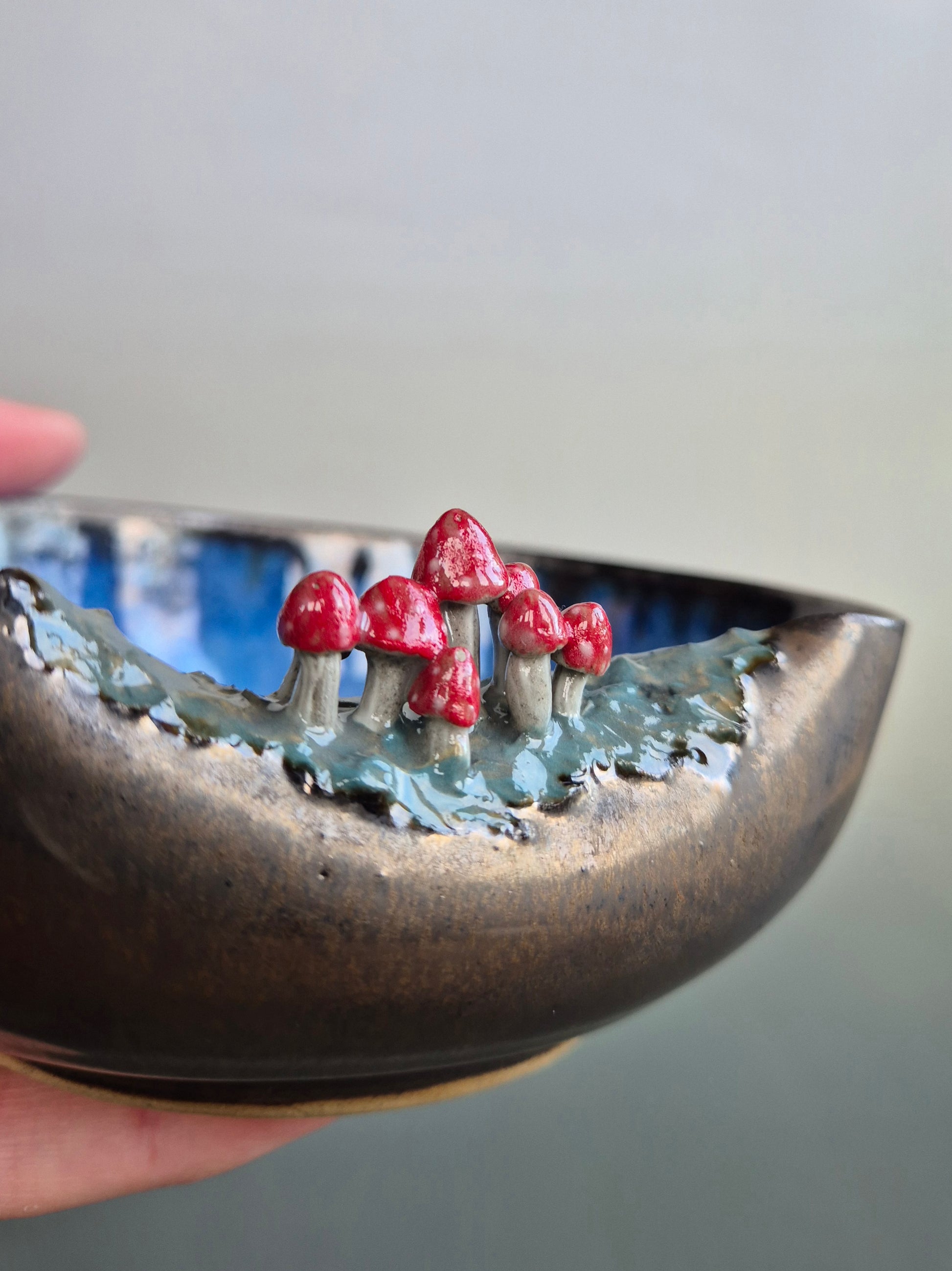 Close up of a small bowl featuring hand sculpted tiny toadstools  on the rim. Bronze glazed exterior. The bowl is resting in a person's hand. On a grey background