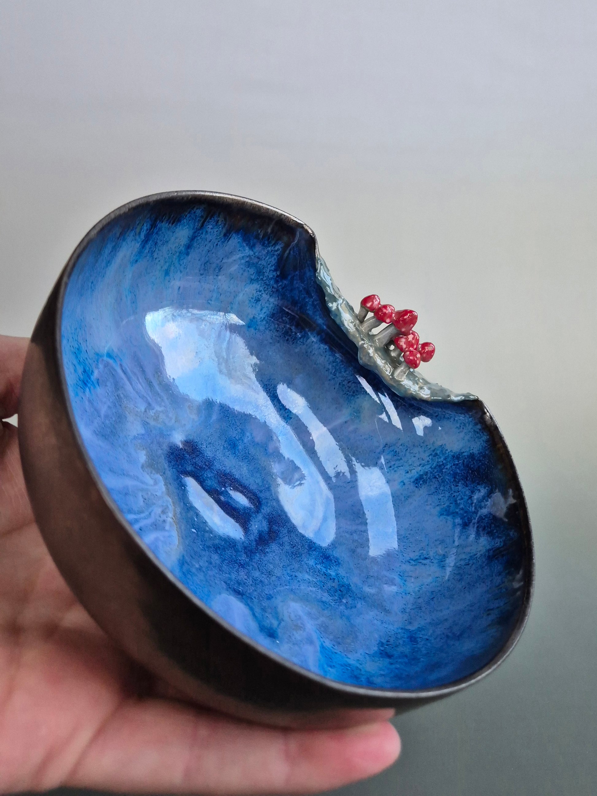 A small bowl featuring hand sculpted tiny toadstools  on the rim. Bronze glazed exterior. The bowl is resting in a person's hand. On a grey background