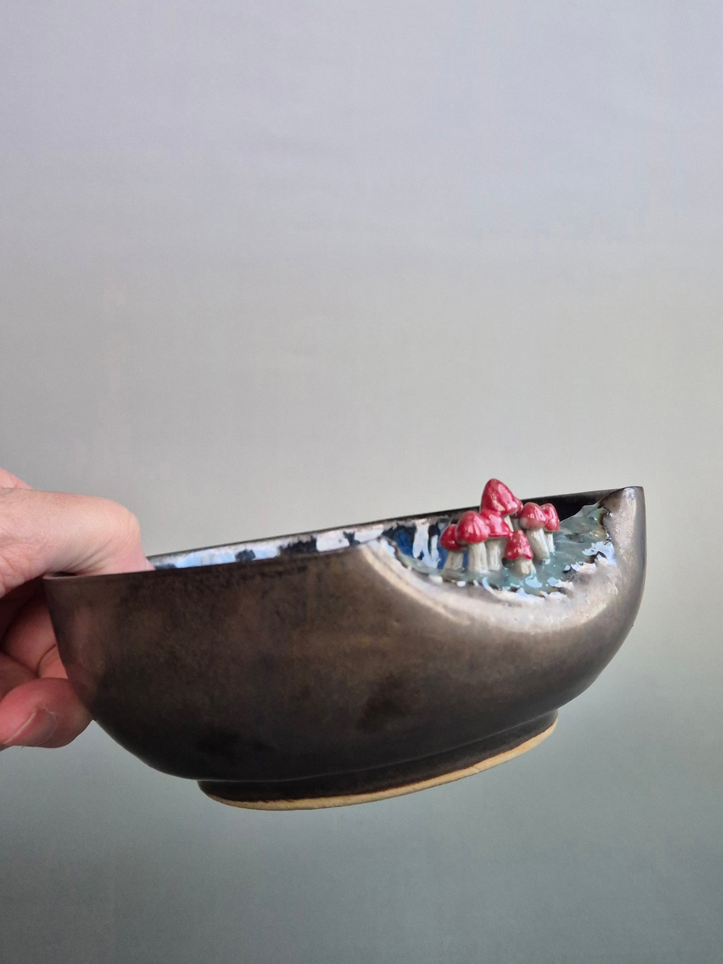 A person holding a small bowl featuring hand sculpted tiny toadstools  on the rim. Bronze glazed exterior. On a grey background