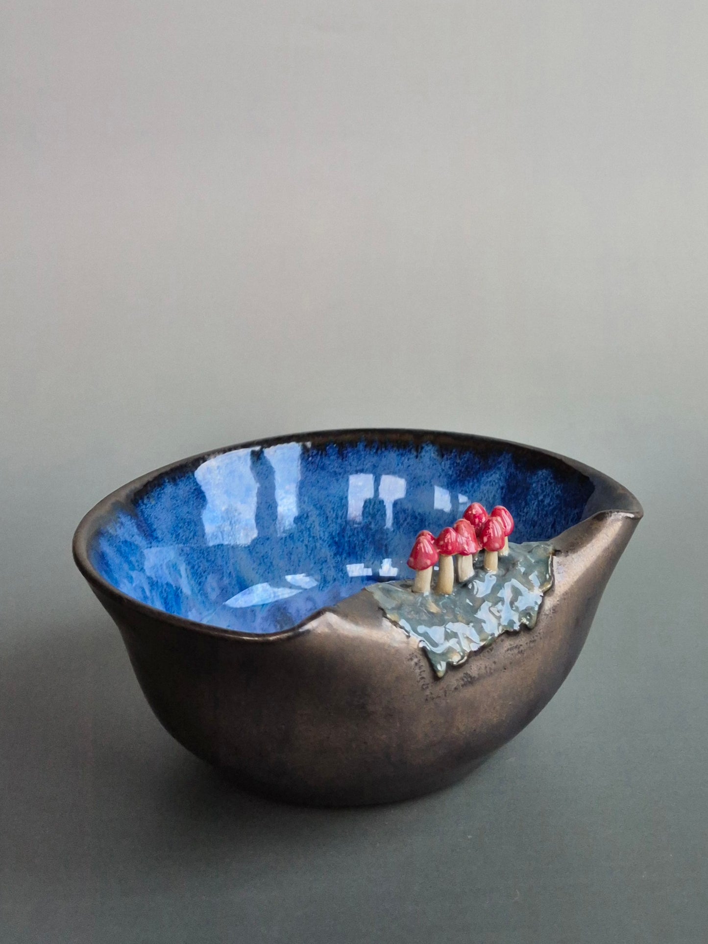 Small trinket dish featuring tiny sculpted toadstools on the rim The bowl has a bronze glazed exterior and a dark blue interior. On a plain background
