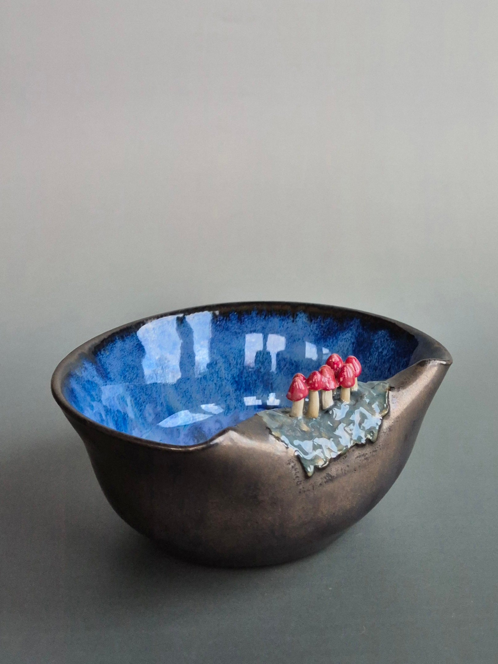 Small trinket dish featuring tiny sculpted toadstools on the rim The bowl has a bronze glazed exterior and a dark blue interior. On a plain background