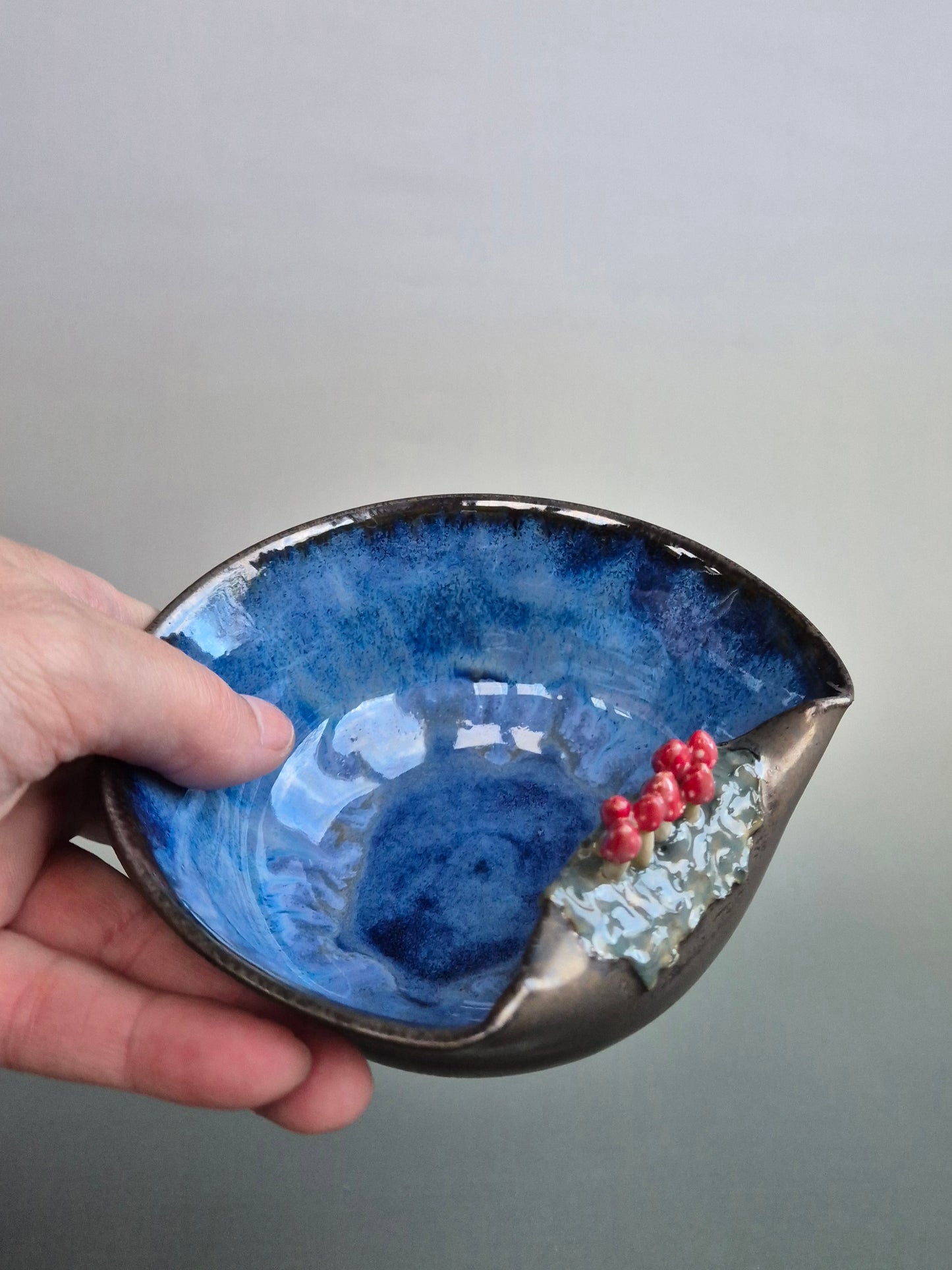 Hand holding a small trinket dish featuring tiny sculpted toadstools on the rim The bowl has a bronze glazed exterior and a dark blue interior. On a plain background