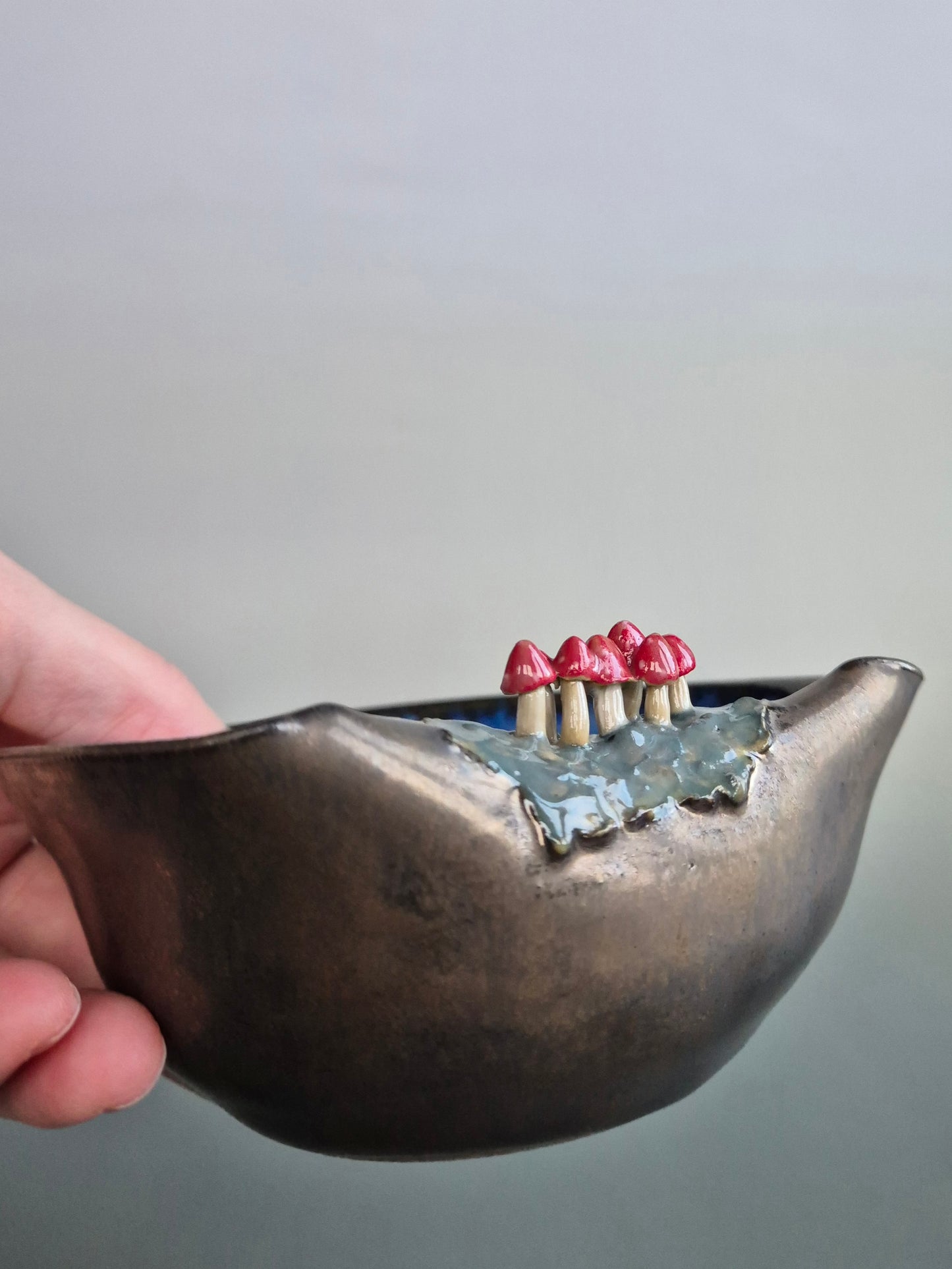 Hand holding a small trinket dish featuring tiny sculpted toadstools on the rim The bowl has a bronze glazed exterior and a dark blue interior. On a plain background
