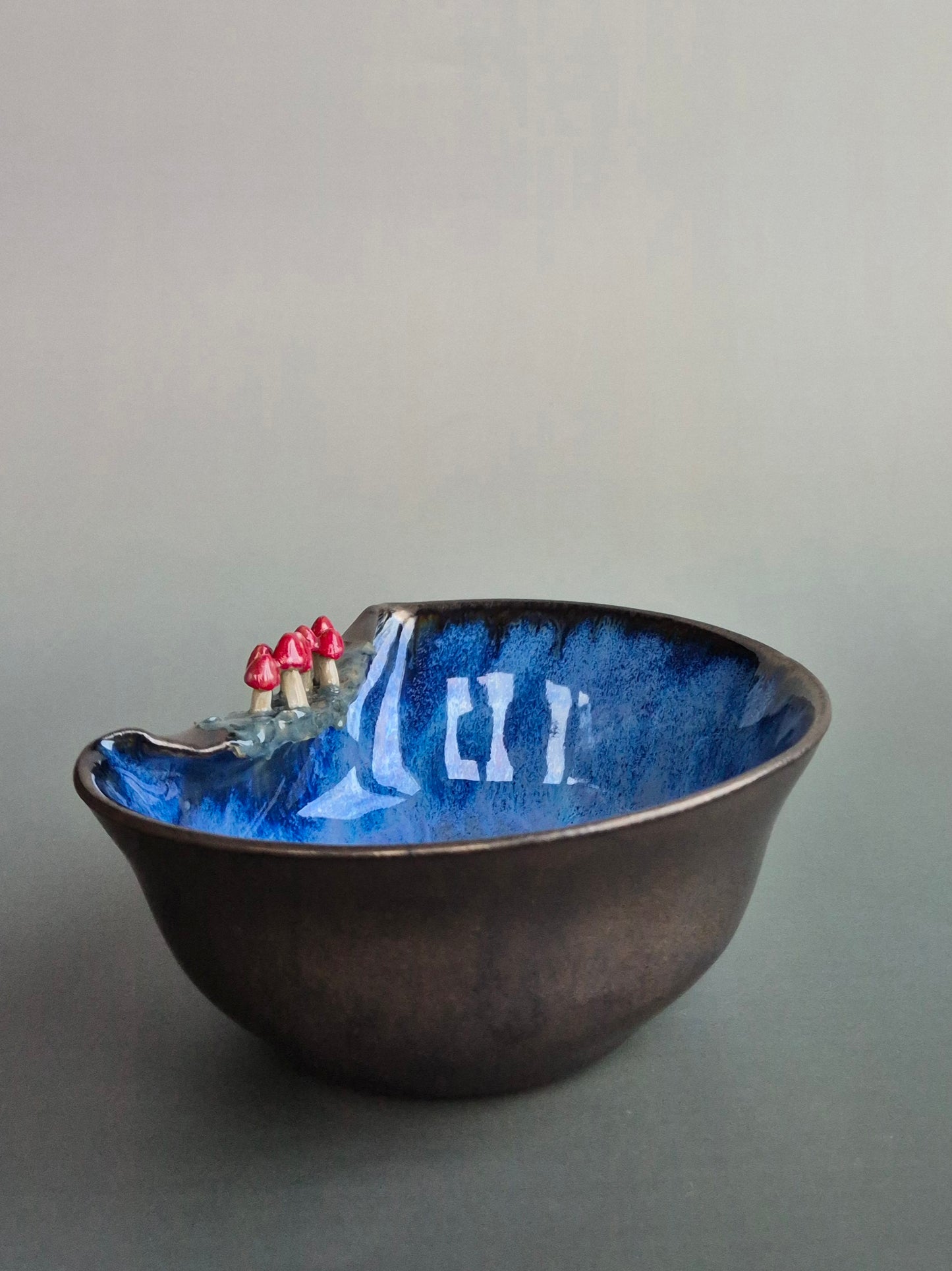A small trinket dish featuring tiny sculpted toadstools on the rim The bowl has a bronze glazed exterior and a dark blue interior. On a plain background