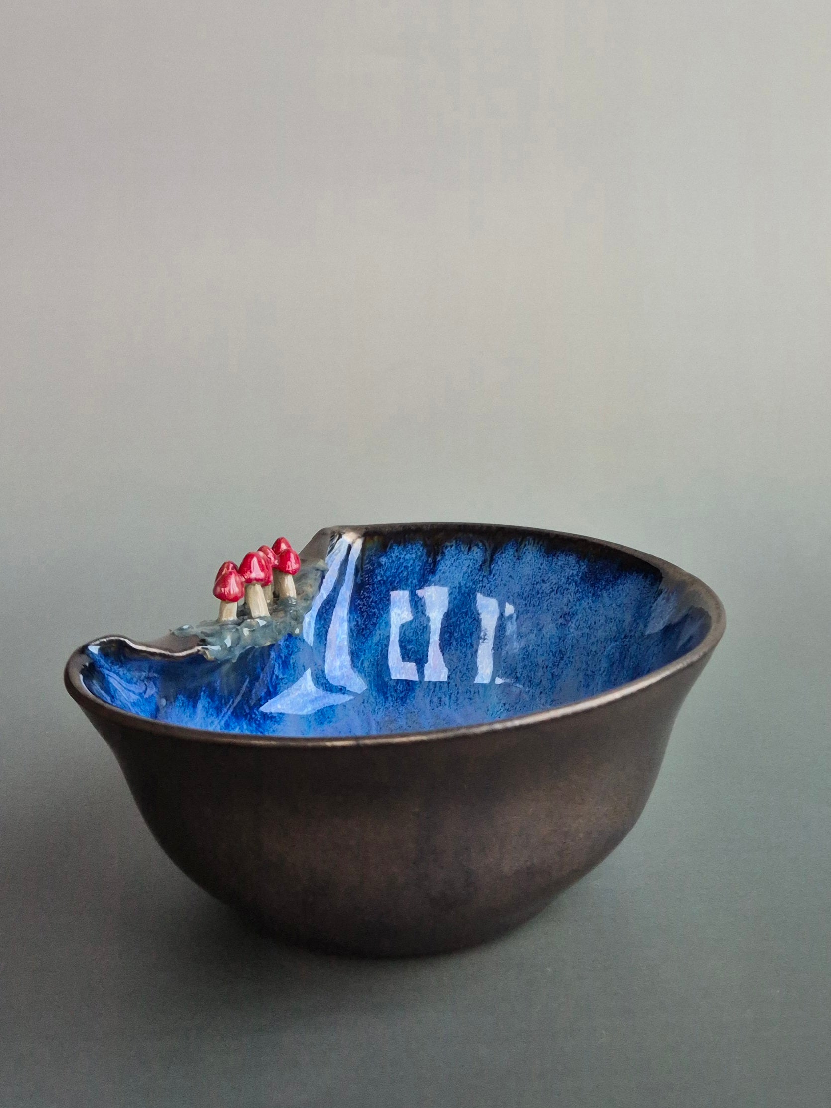 A small trinket dish featuring tiny sculpted toadstools on the rim The bowl has a bronze glazed exterior and a dark blue interior. On a plain background