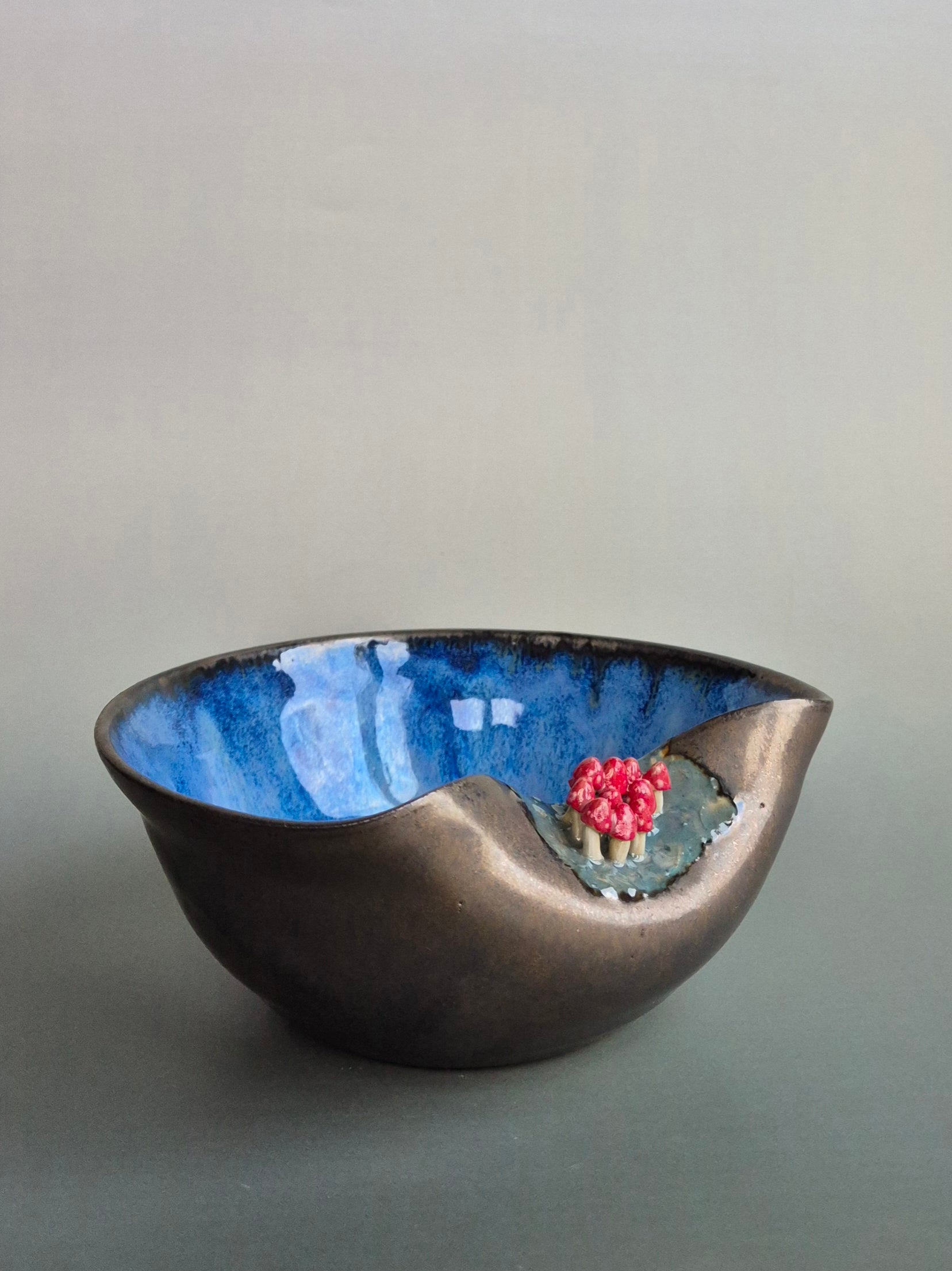 Small trinket dish featuring tiny hand sculpted toadstools on the rim. Dish has a bronze lazed exterior and a deep blue interior. On a plain background