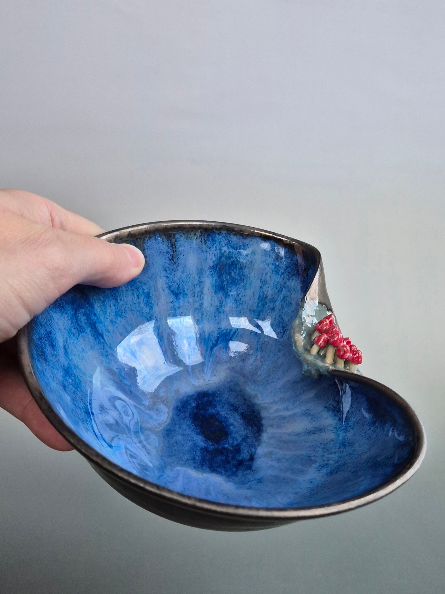 A hand holding a small trinket dish featuring tiny hand sculpted toadstools on the rim. Dish has a bronze lazed exterior and a deep blue interior. On a plain background