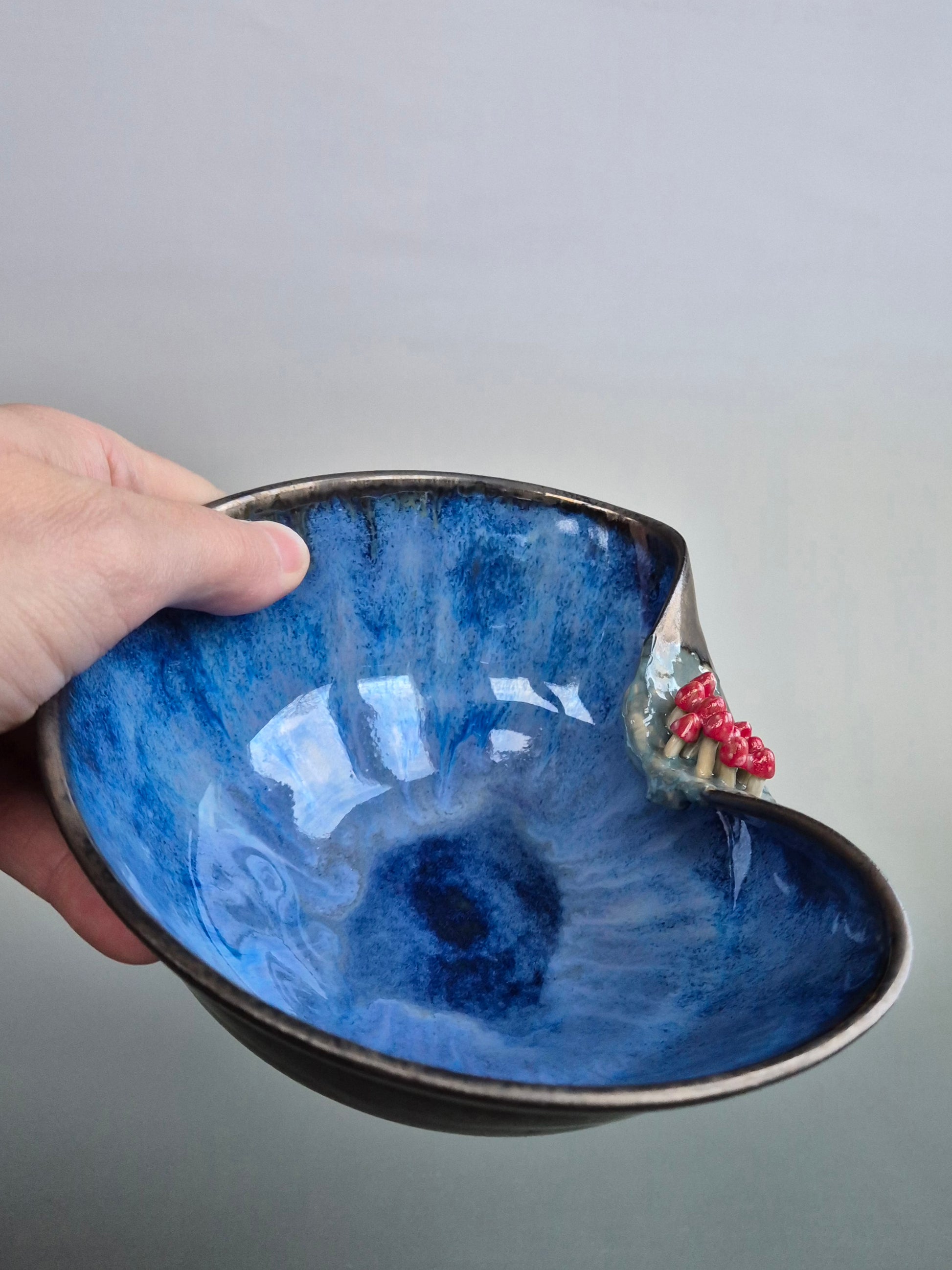 A hand holding a small trinket dish featuring tiny hand sculpted toadstools on the rim. Dish has a bronze lazed exterior and a deep blue interior. On a plain background