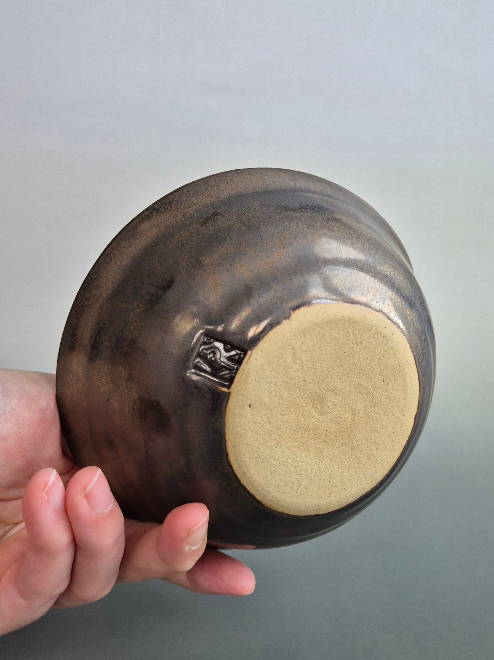 A hand holding a small trinket dish to show the plain base with the makers mark. Dish has a bronze glazed exterior. On a plain background