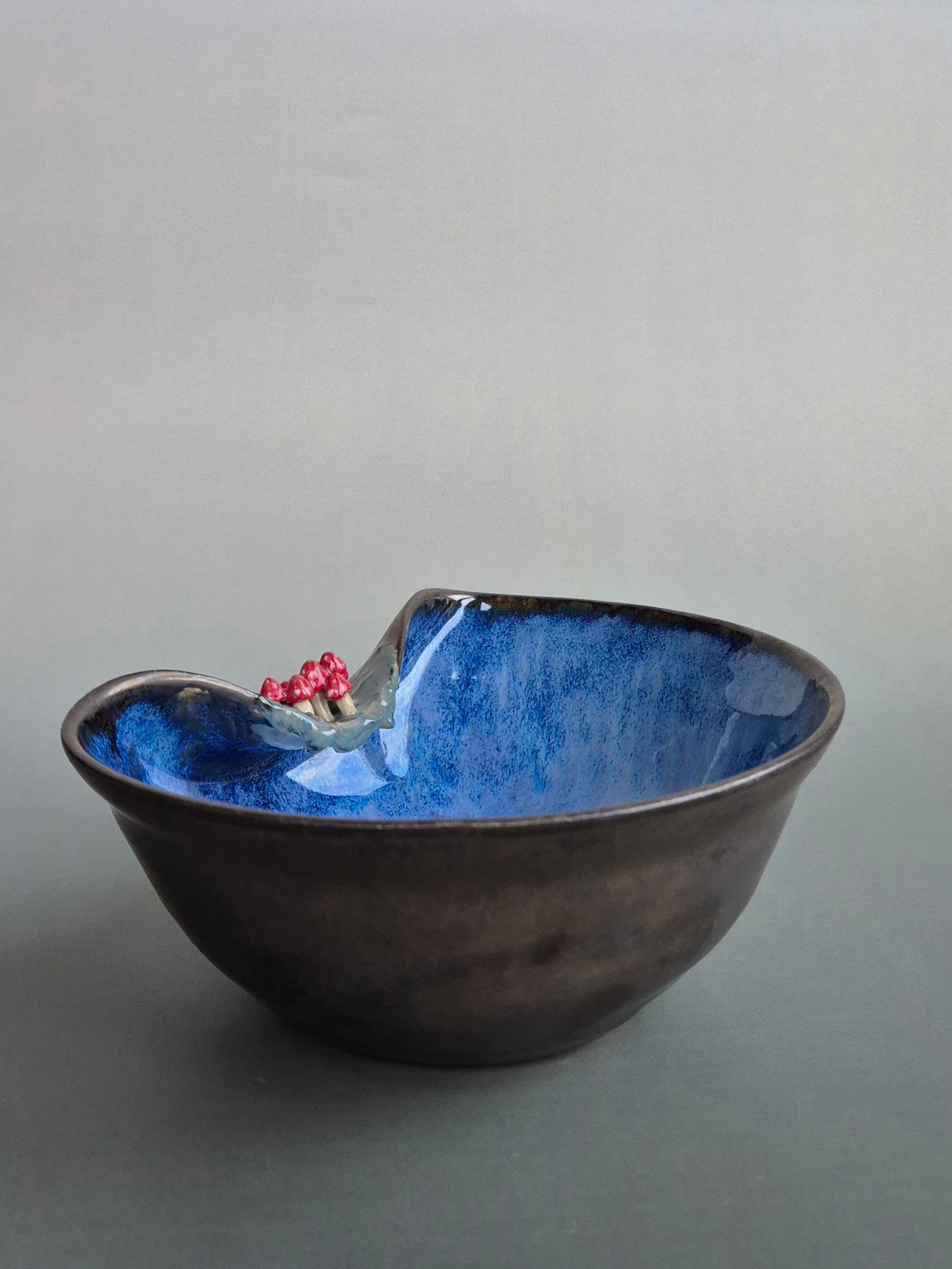 Small trinket dish featuring tiny hand sculpted toadstools on the rim. Dish has a bronze lazed exterior and a deep blue interior. On a plain background
