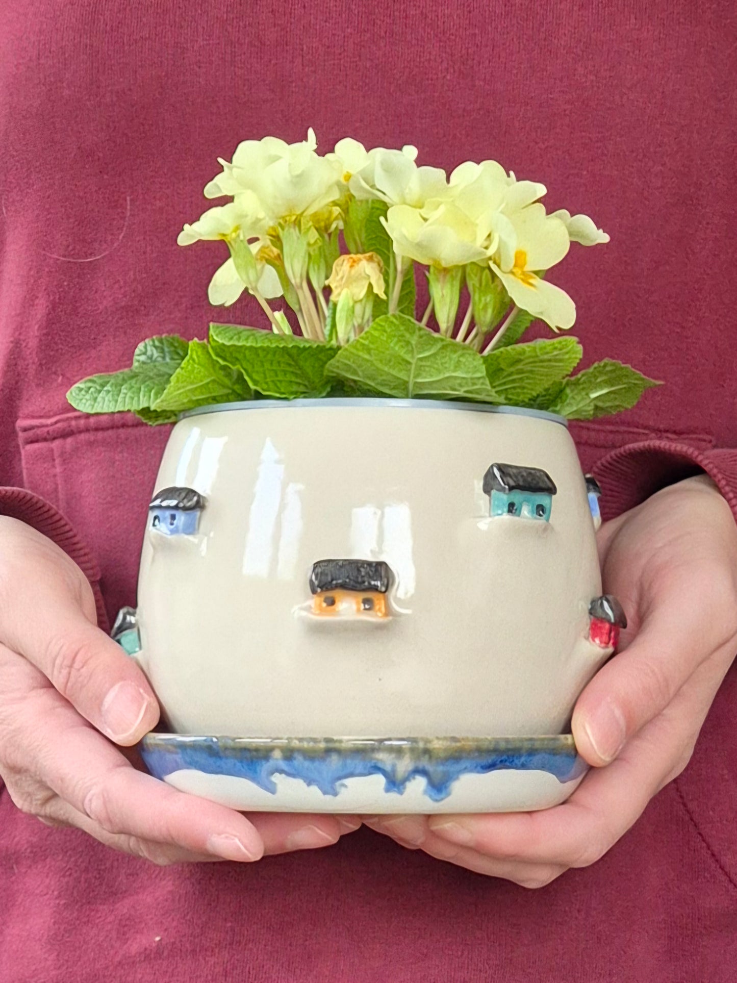  person holding a Ceramic planter decorated with colourful tiny houses. On a blue saucer with a plain background.