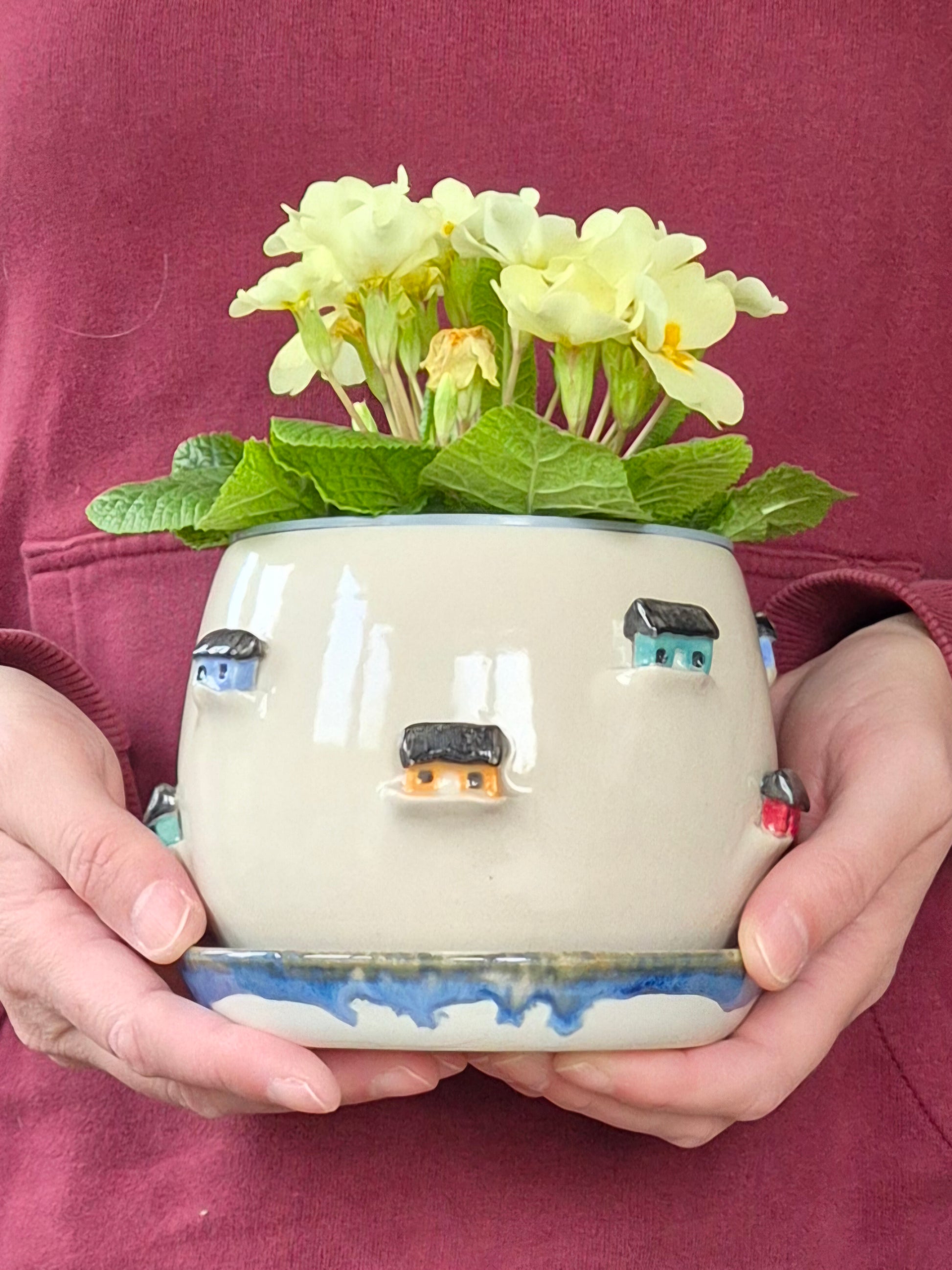  person holding a Ceramic planter decorated with colourful tiny houses. On a blue saucer with a plain background.