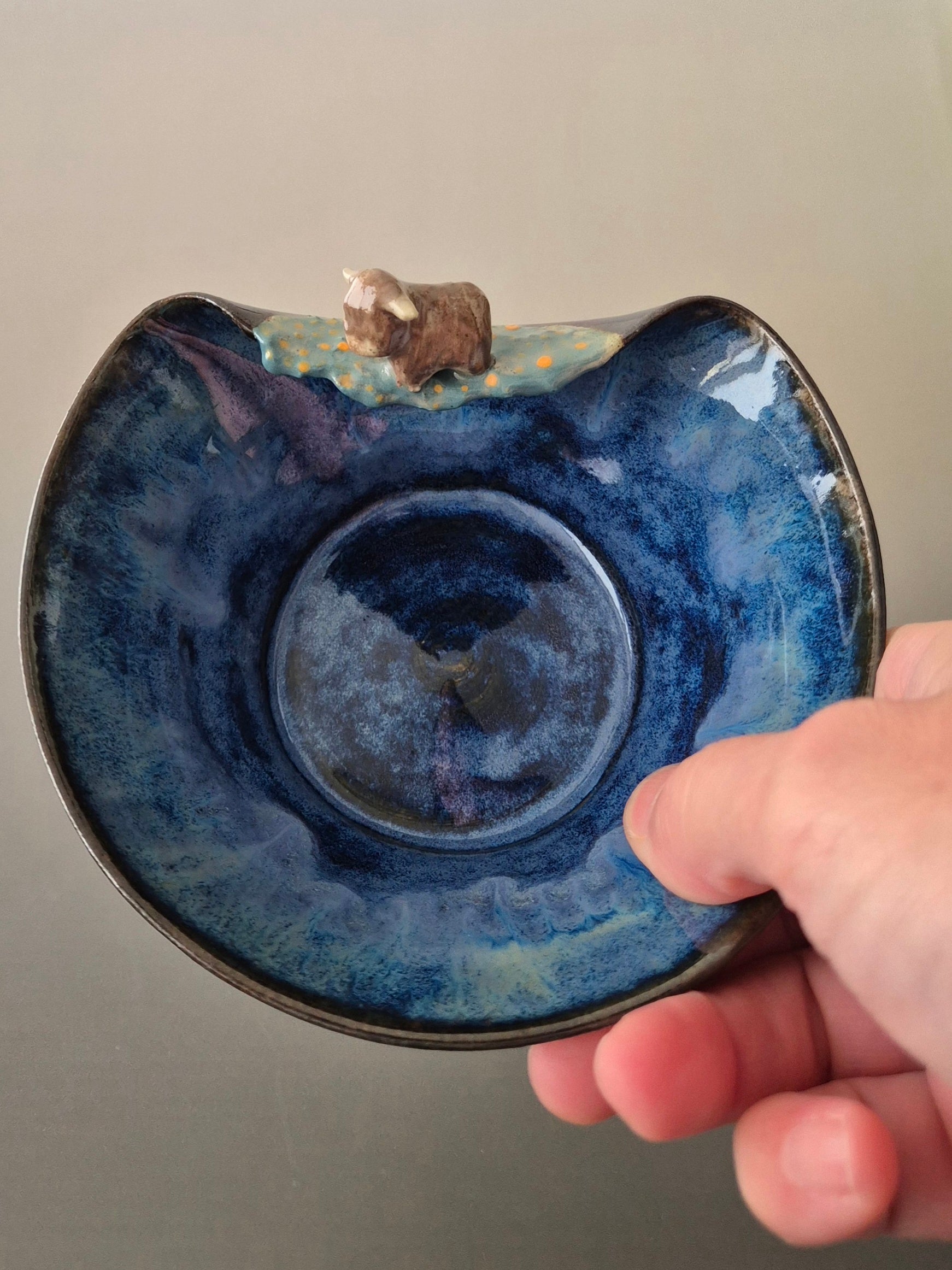 A hand holding a small ceramic bowl with a tiny sculpted highland cow. On a grey background