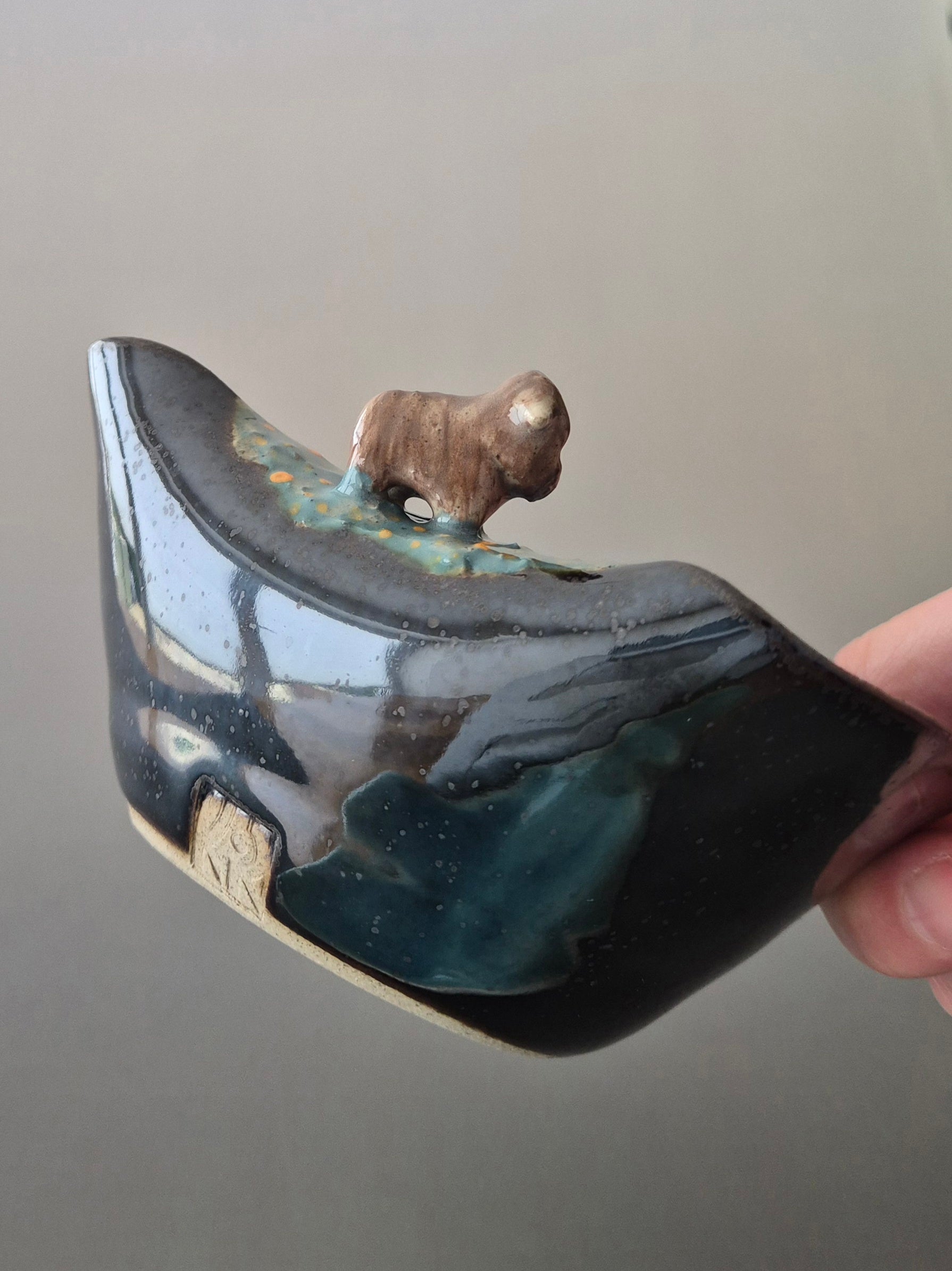 A hand holding a small ceramic bowl with a tiny sculpted highland cow. On a grey background