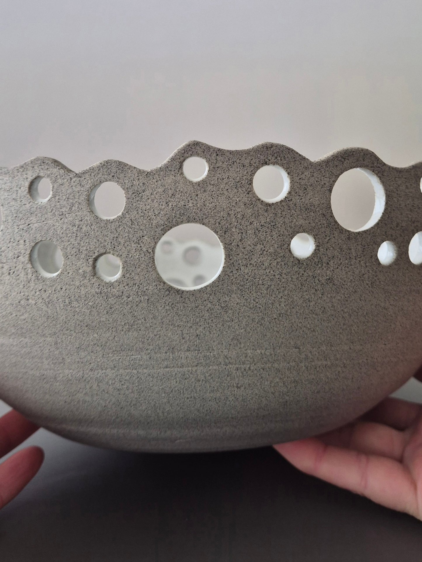Ceramic bowl with decorative holes held by a hand against a neutral background