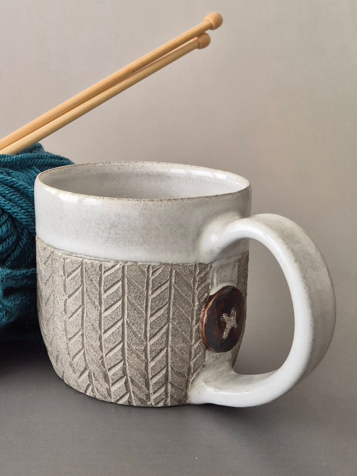 Gray textured mug with white handle next to yarn and knitting needles on a gray background