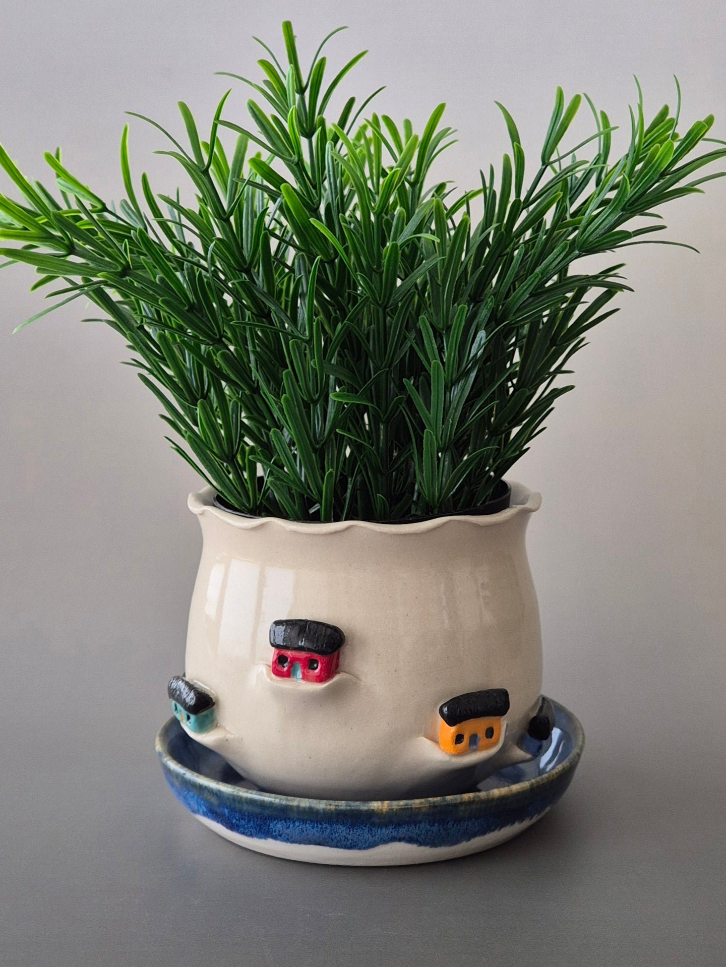Ceramic plant pot with a green plant. Pot is decorated with tiny houses on a blue glazed saucer. With a plain background