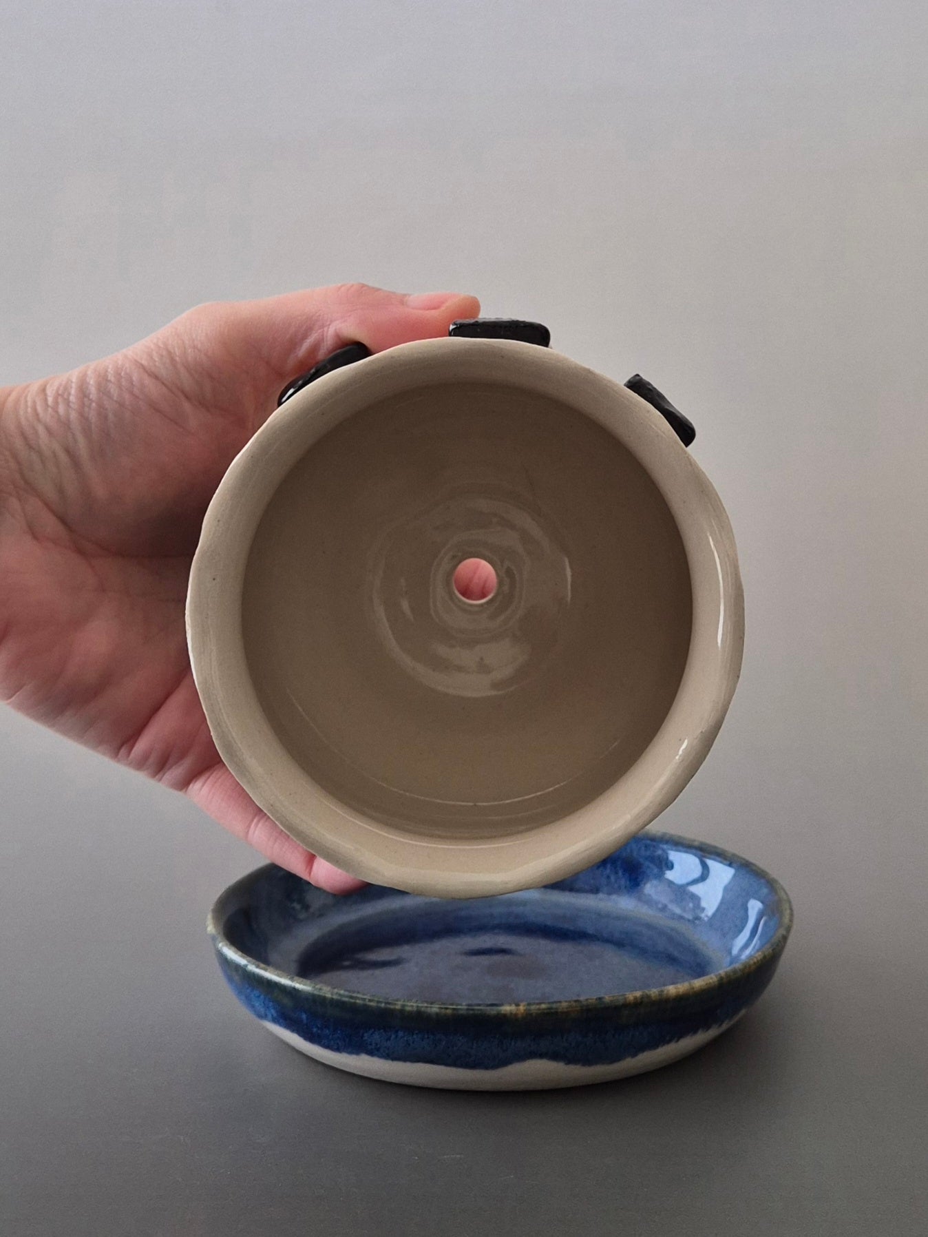 Hand holding a ceramic plant pot to show the inside and drainage hole above a blue glazed saucer. With a plain background. 