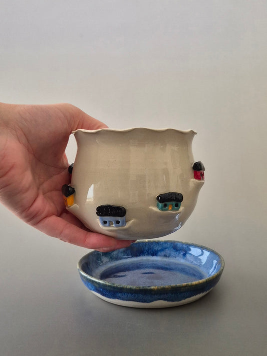 Hand holding a ceramic plant pot decorated with tiny houses above a blue glazed saucer. With a plain background
