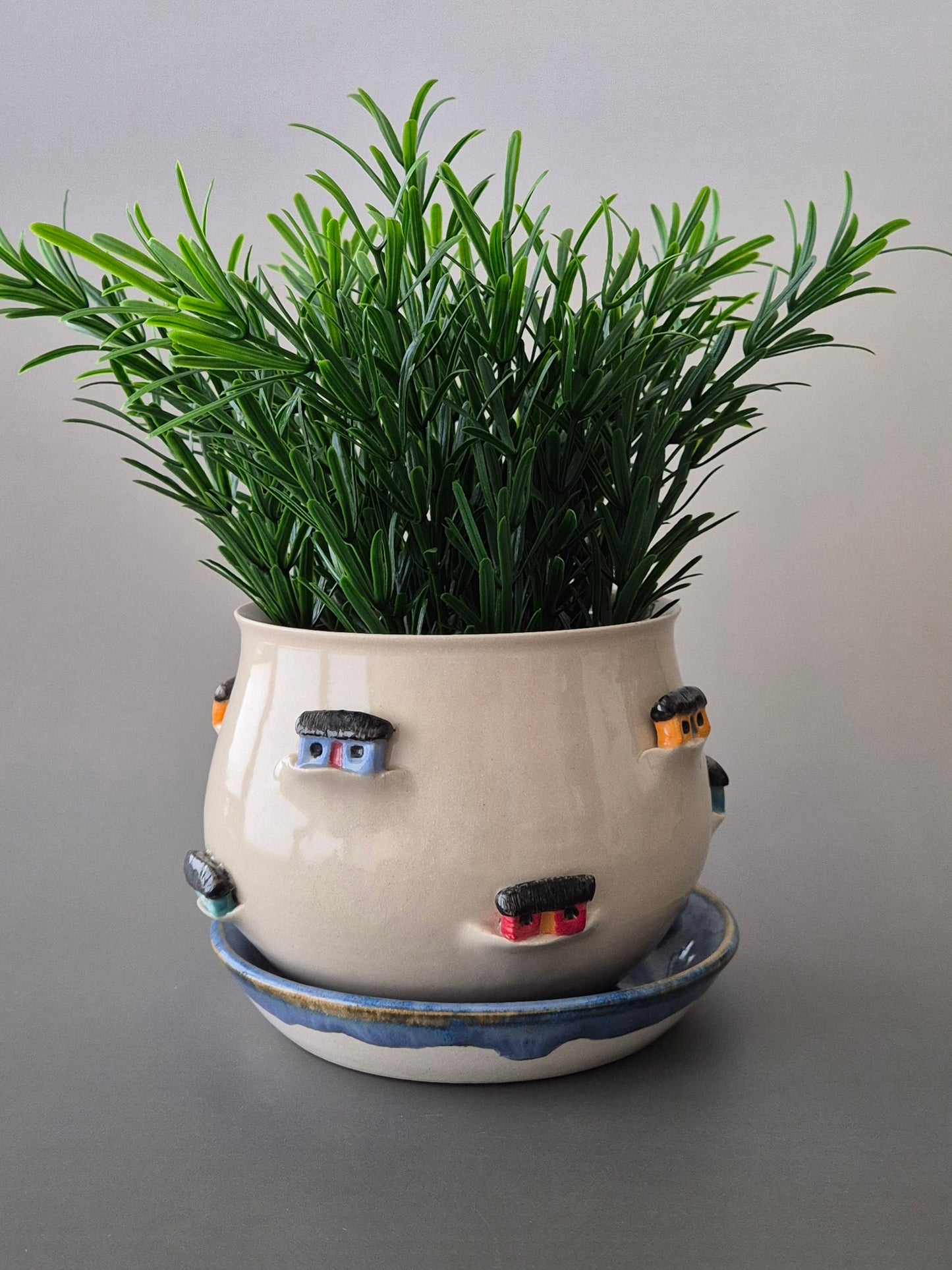 Ceramic plant pot with colourful tiny houses around the outer edge. Containing a green plant on a gray background