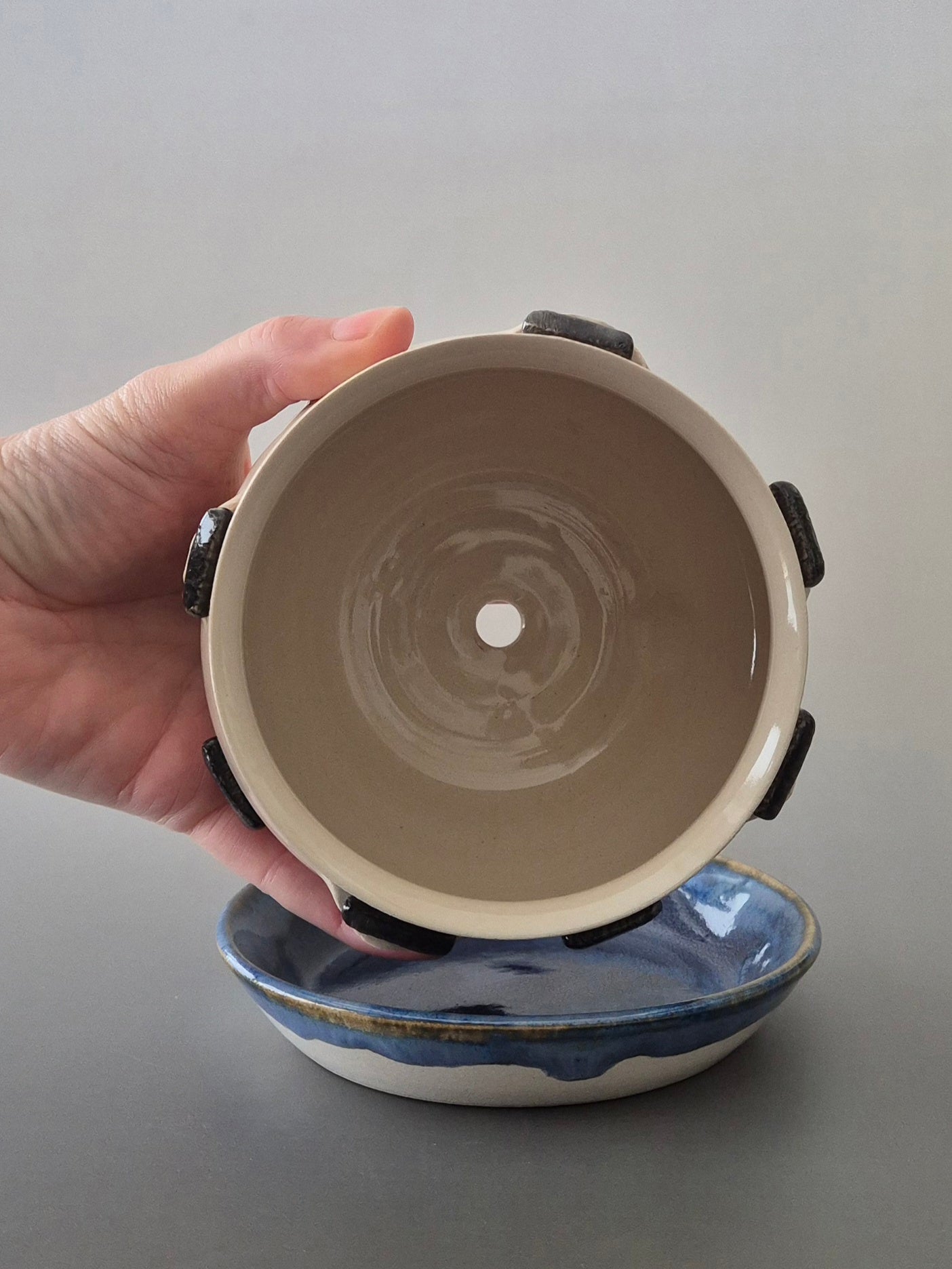 Hand holding a ceramic plant pot to show the inside and drainage hole with a saucer underneath. On a gray background