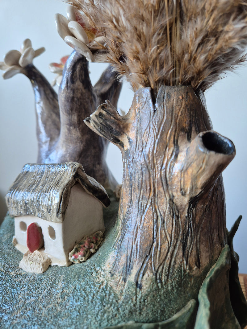Decorative sculpture of a small house with a flowering tree behind it and a tree trunk that is also a vase in the front. On a white background