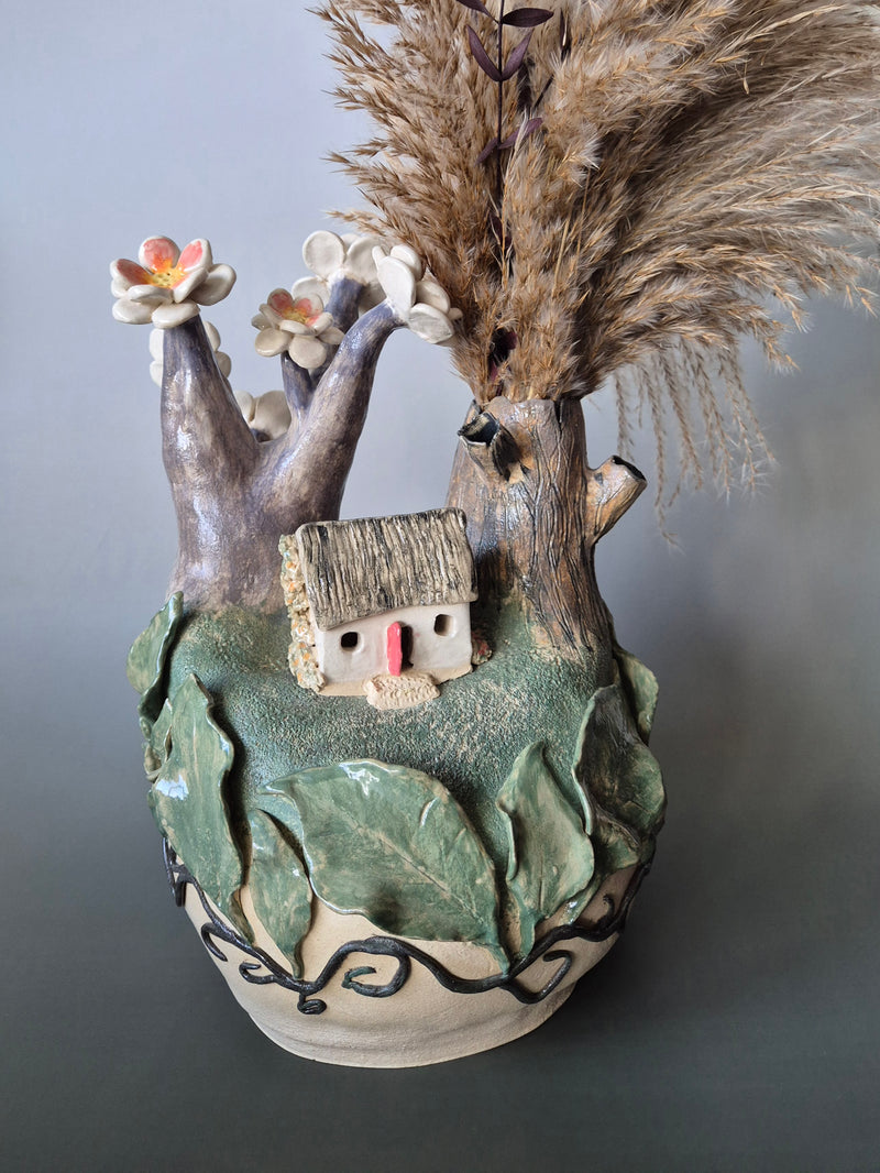 Ceramic vase with decorative elements including a small house, flowers, and natural materials on a gray background.