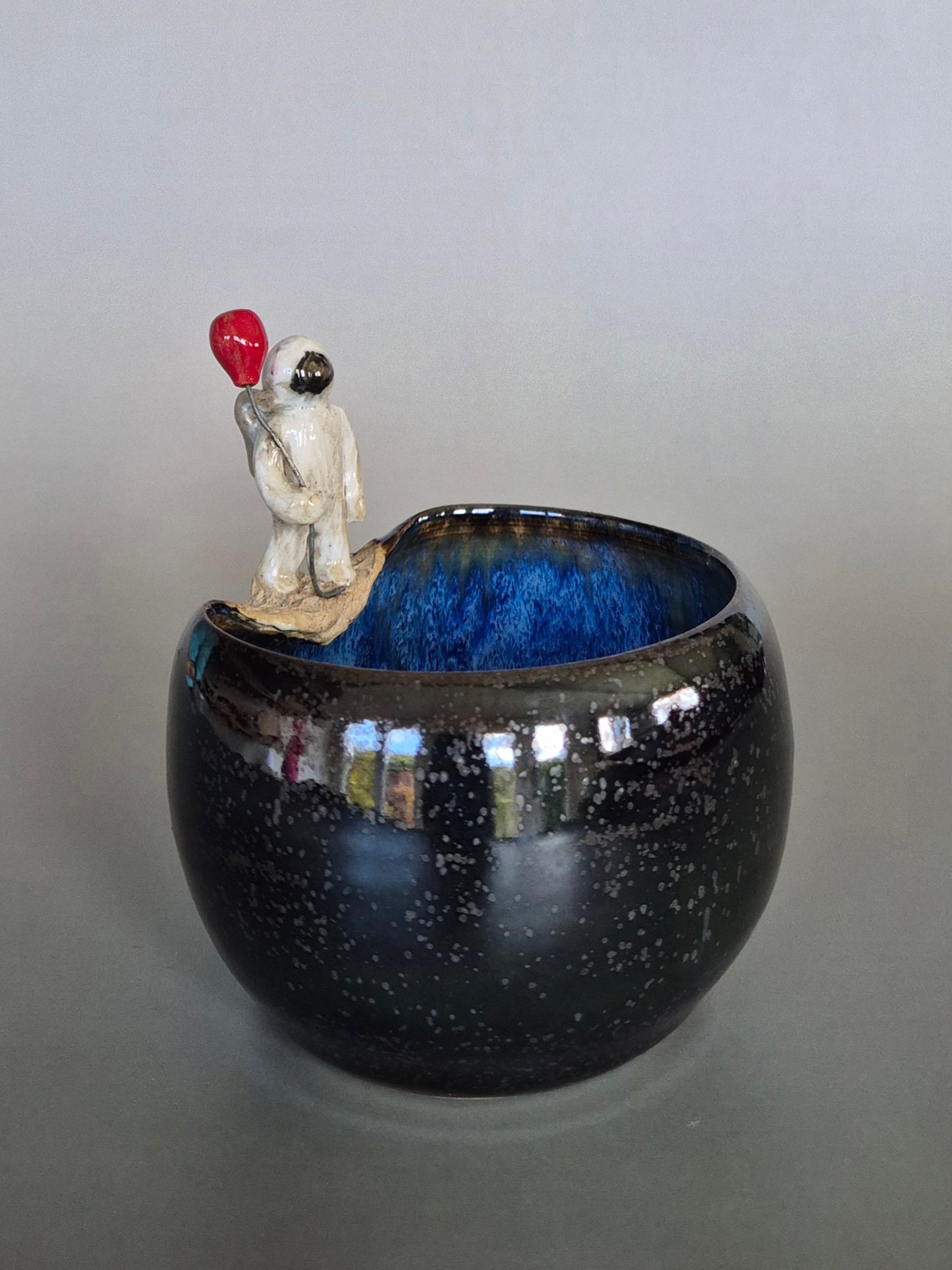 Ceramic bowl with a small sculptured figurine of an astronaut holding a red balloon. On a plain background