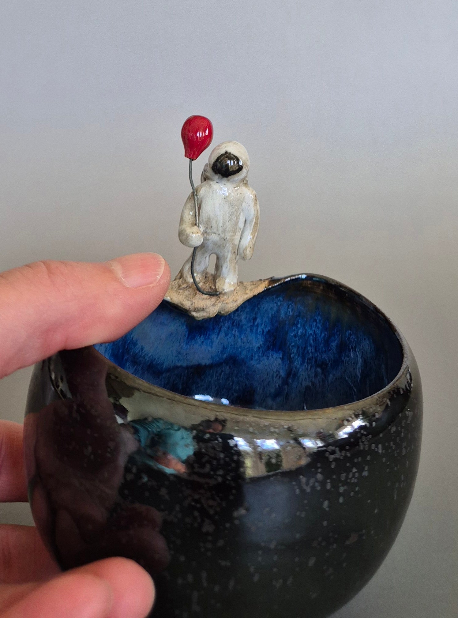 A person's hand holding a ceramic bowl with a small sculptured figurine of an astronaut holding a red balloon. On a plain background