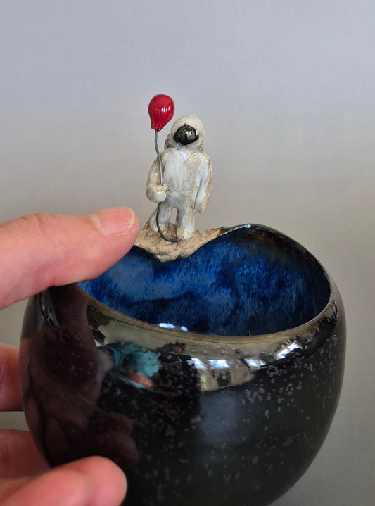 A person's hand holding a ceramic bowl with a small sculptured figurine of an astronaut holding a red balloon. On a plain background