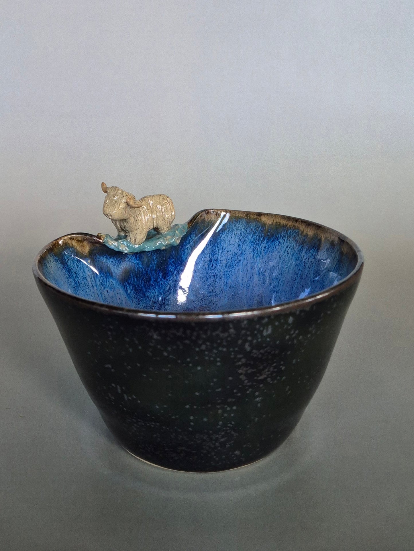 A small ceramic bowl featuring a tiny sculpted figure of a highland cow. On a plain grey background.