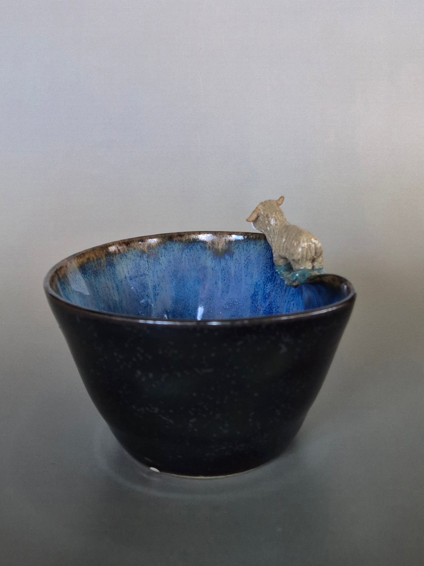 A small ceramic bowl featuring a tiny sculpted figure of a highland cow. On a plain grey background.