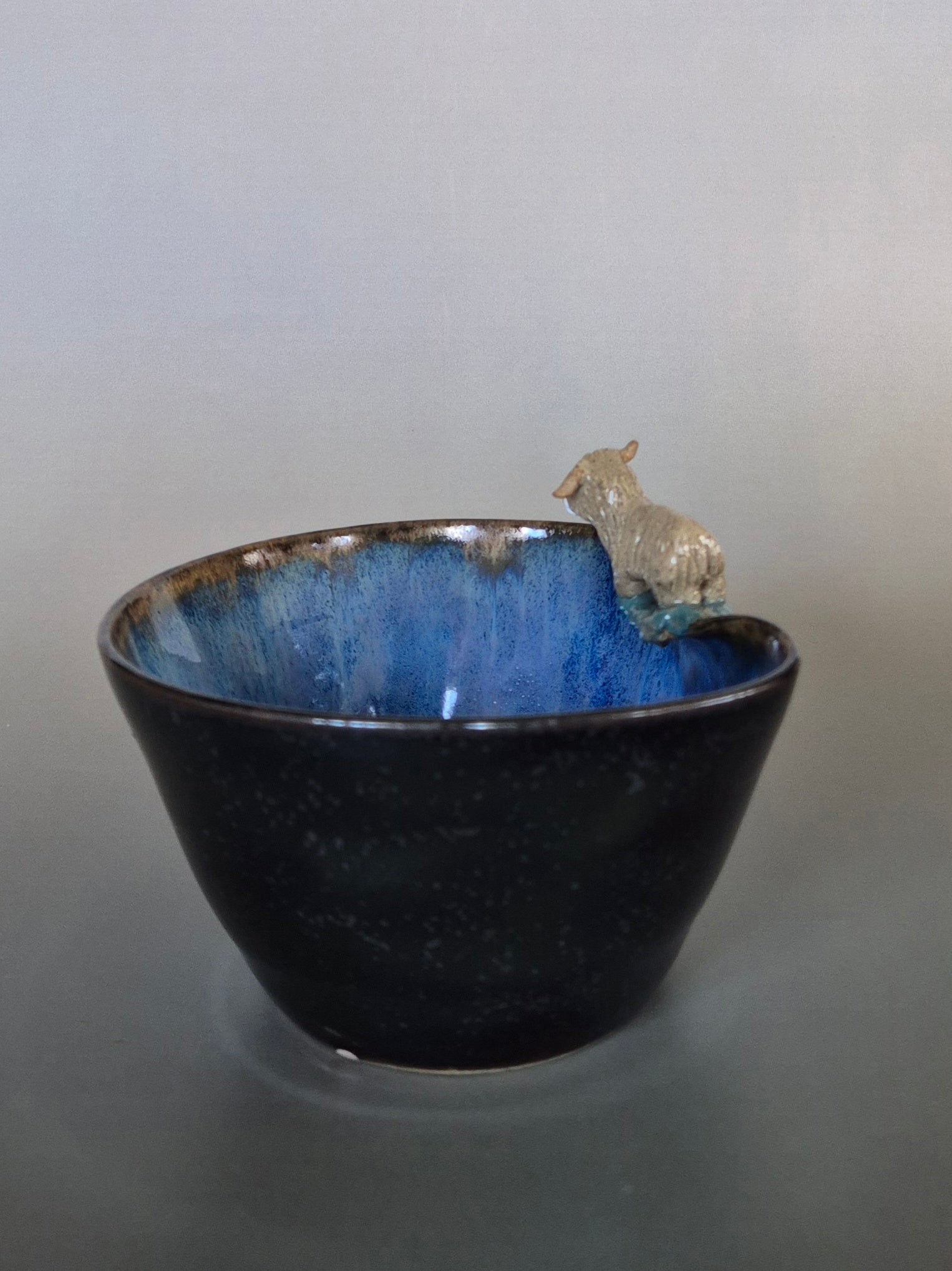 A small ceramic bowl featuring a tiny sculpted figure of a highland cow. On a plain grey background.