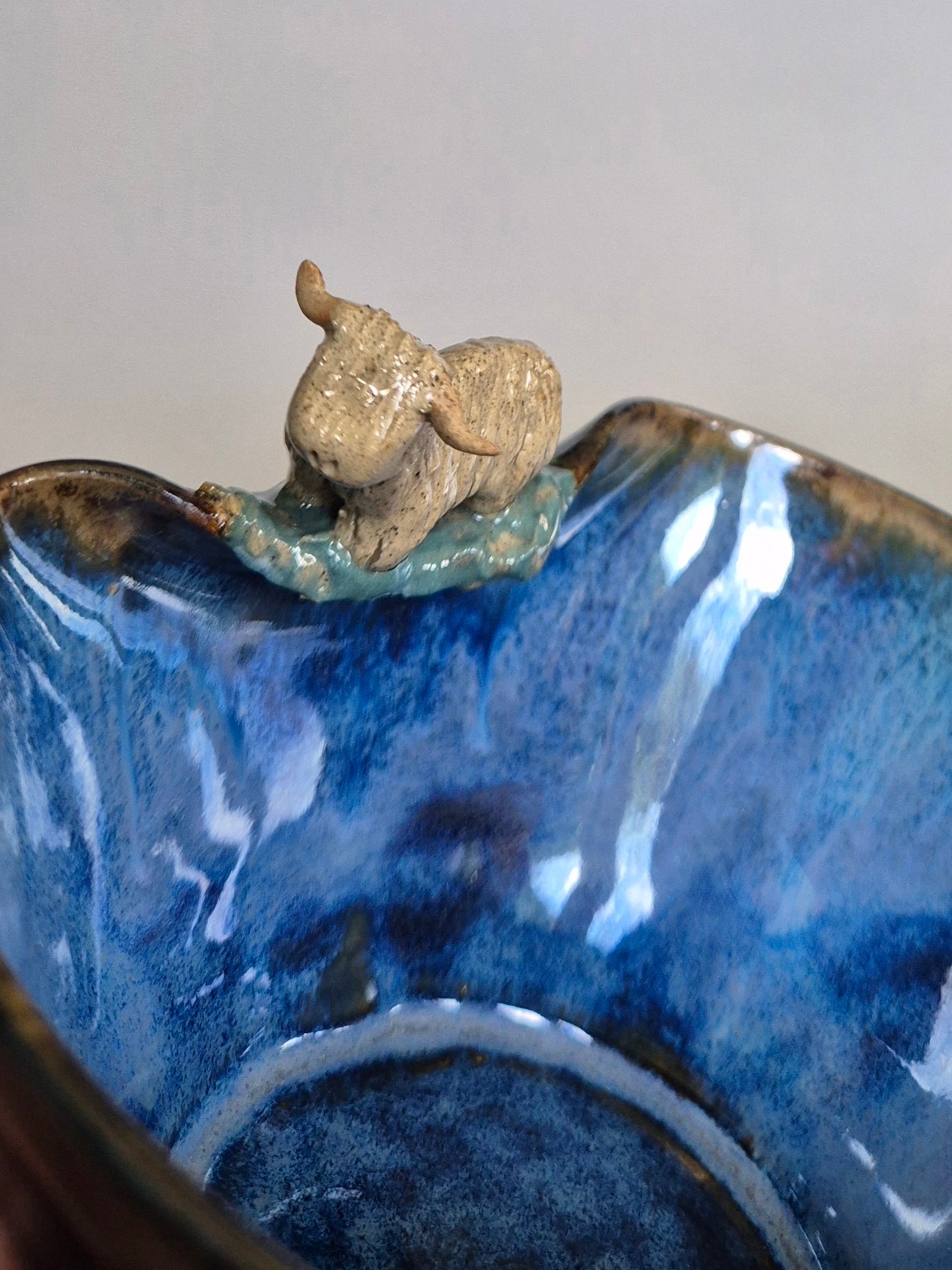 A small ceramic bowl featuring a tiny sculpted figure of a highland cow. On a plain grey background.