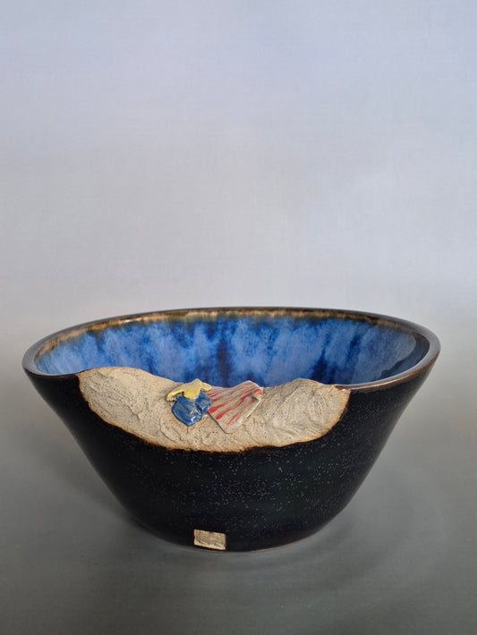 A ceramic bowl featuring sculptural details creating a miniature beach scene. On a plain background