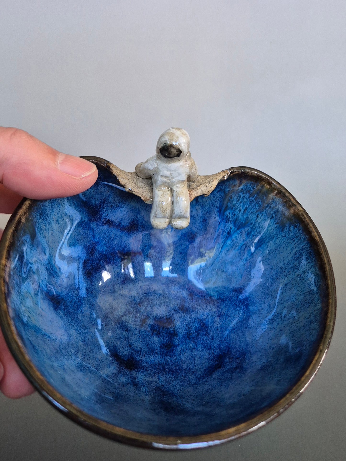 Hand holding a bowl decorated with a small figurine of an astronaut sitting on the edge. On a plain background