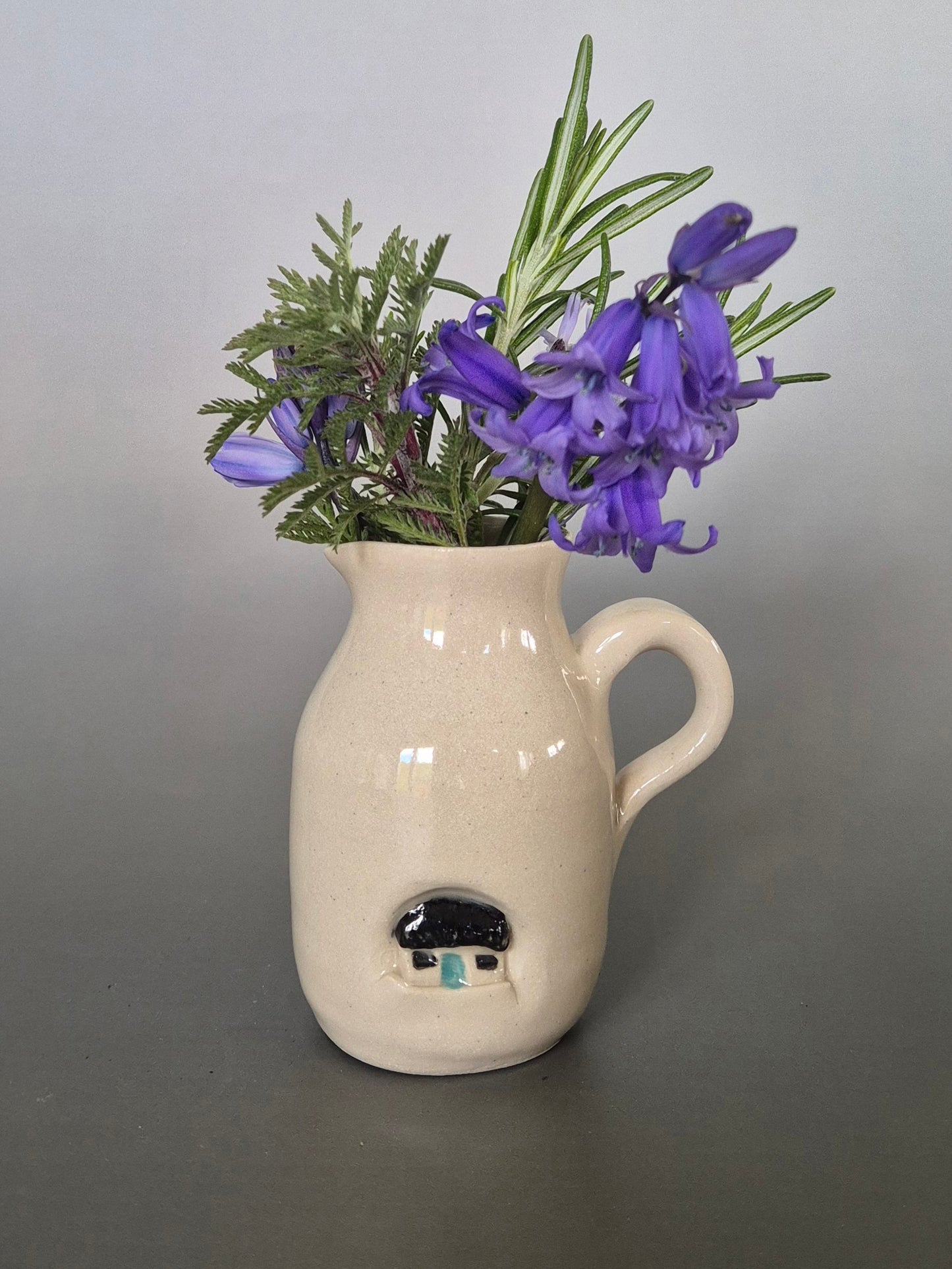 White ceramic vase with a small house design on a gray background, filled with purple flowers and green leaves.
