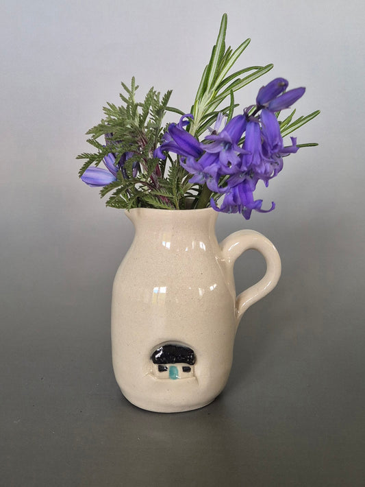 White ceramic vase with a small house design on a gray background, filled with purple flowers and green leaves.
