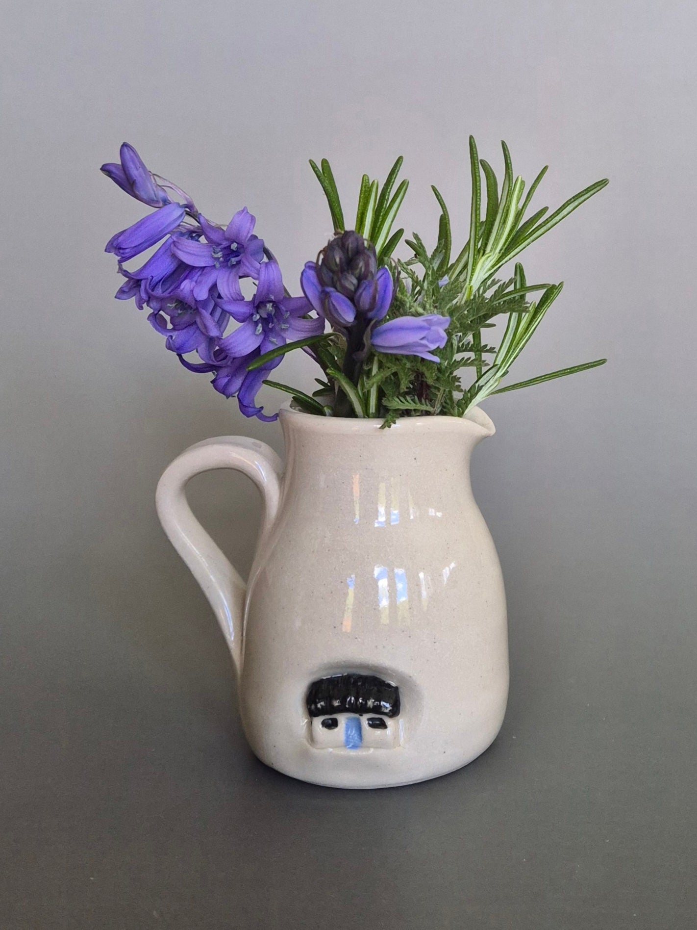 White ceramic pitcher with house holding purple flowers on a gray background