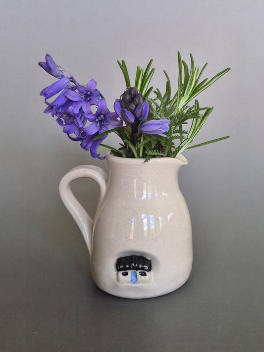White ceramic pitcher with house holding purple flowers on a gray background