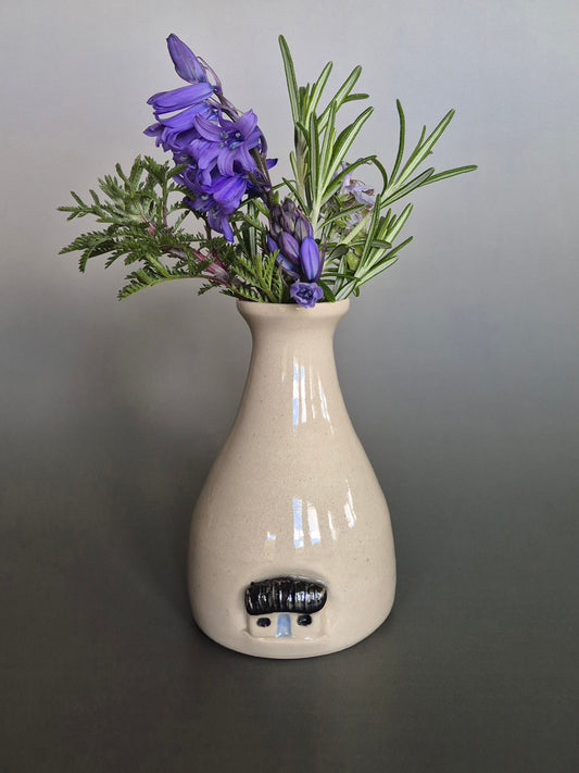 Small white ceramic bottle or bud vase decorated with a small house and filled with wildflowers. On a plain background