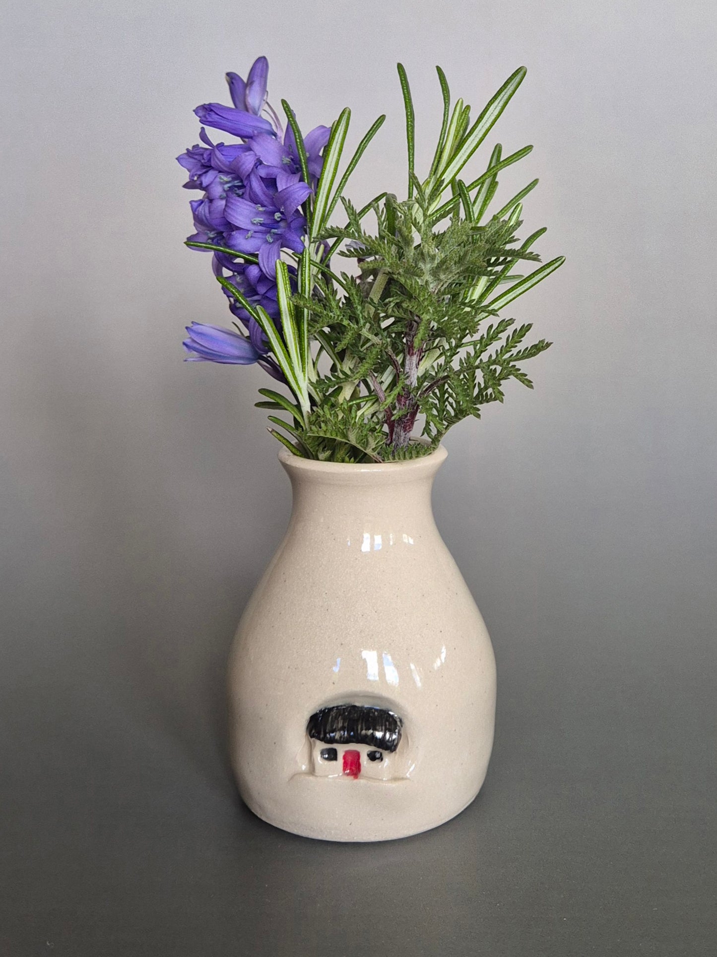 Small white ceramic bottle or bud vase decorated with a small house and filled with wildflowers. On a plain background