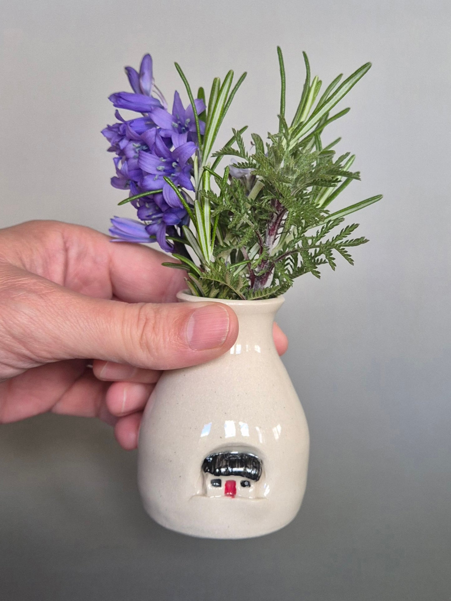 Hand holding a small white ceramic bottle with herbs and flowers inside against a gray background
