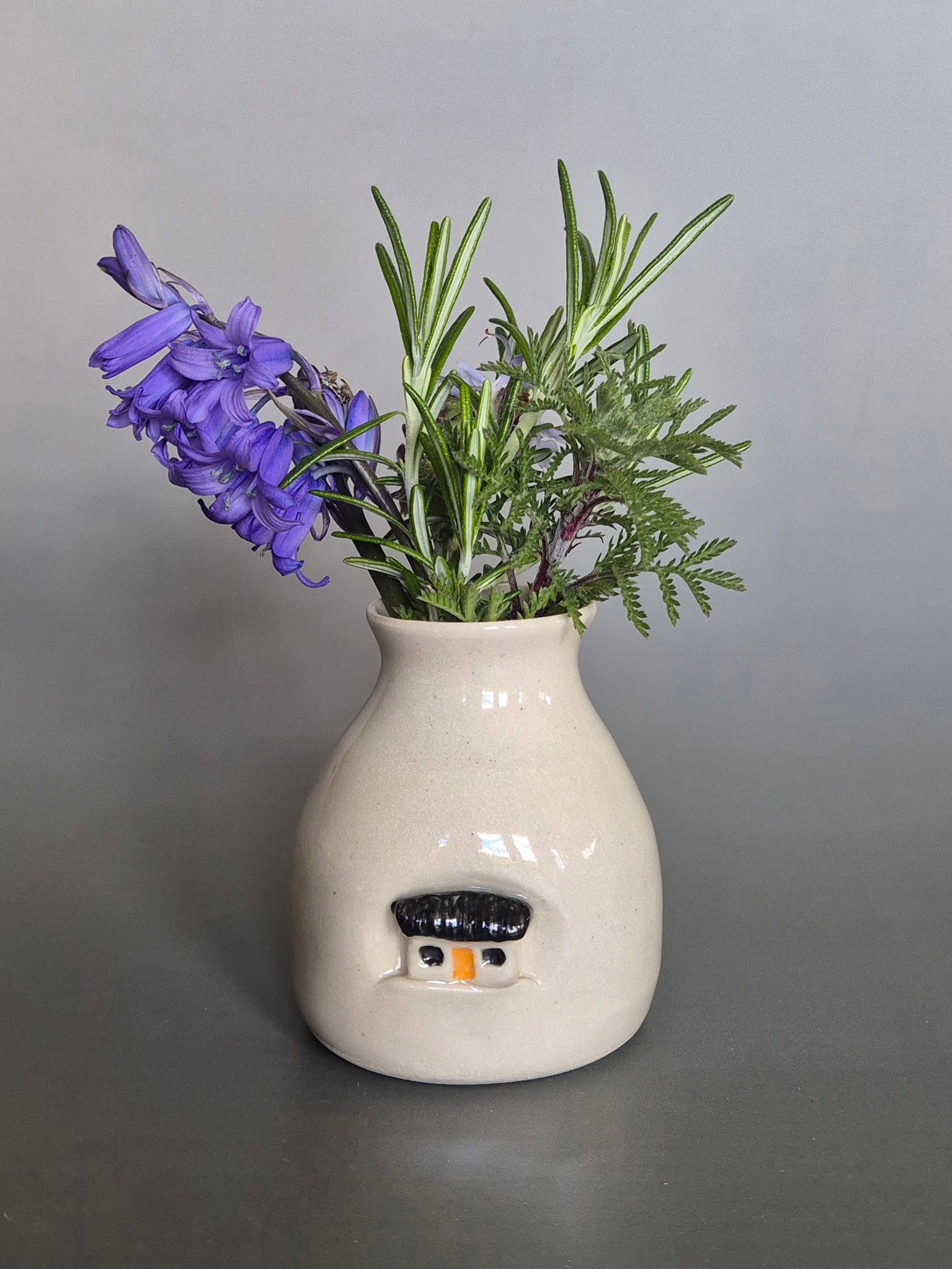Small white ceramic bottle or bud vase decorated with a small house and filled with wildflowers. On a plain background