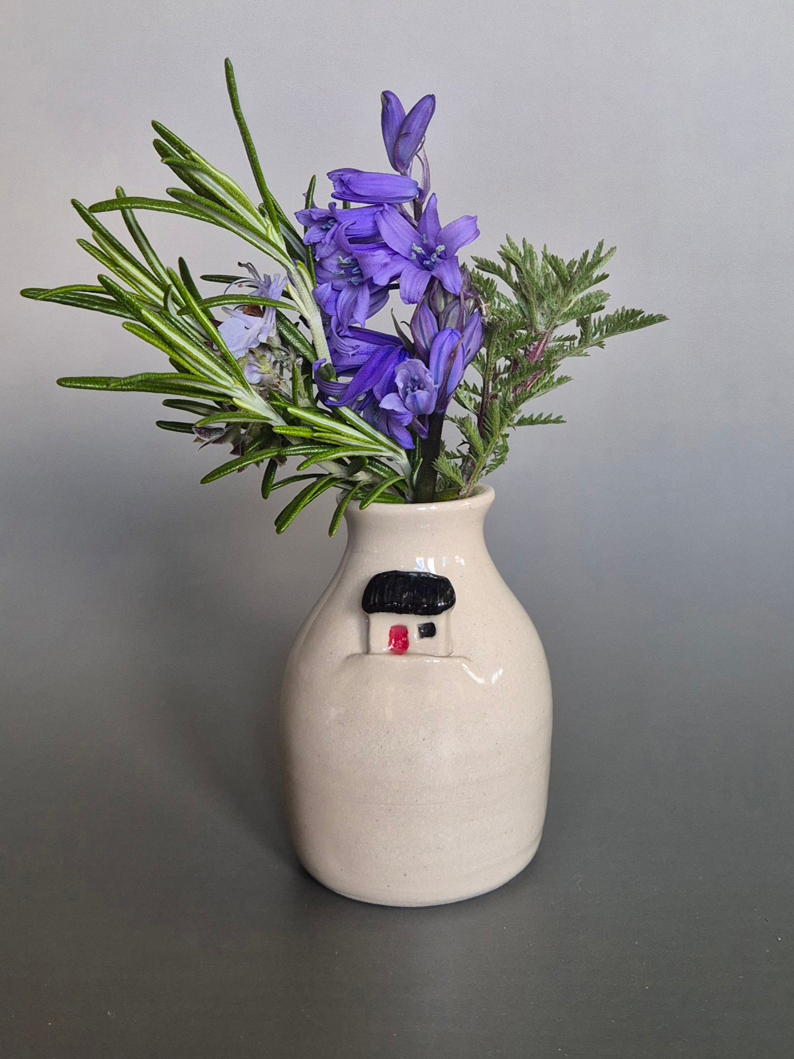 Small white ceramic bottle or bud vase decorated with a small house and filled with wildflowers. On a plain background.