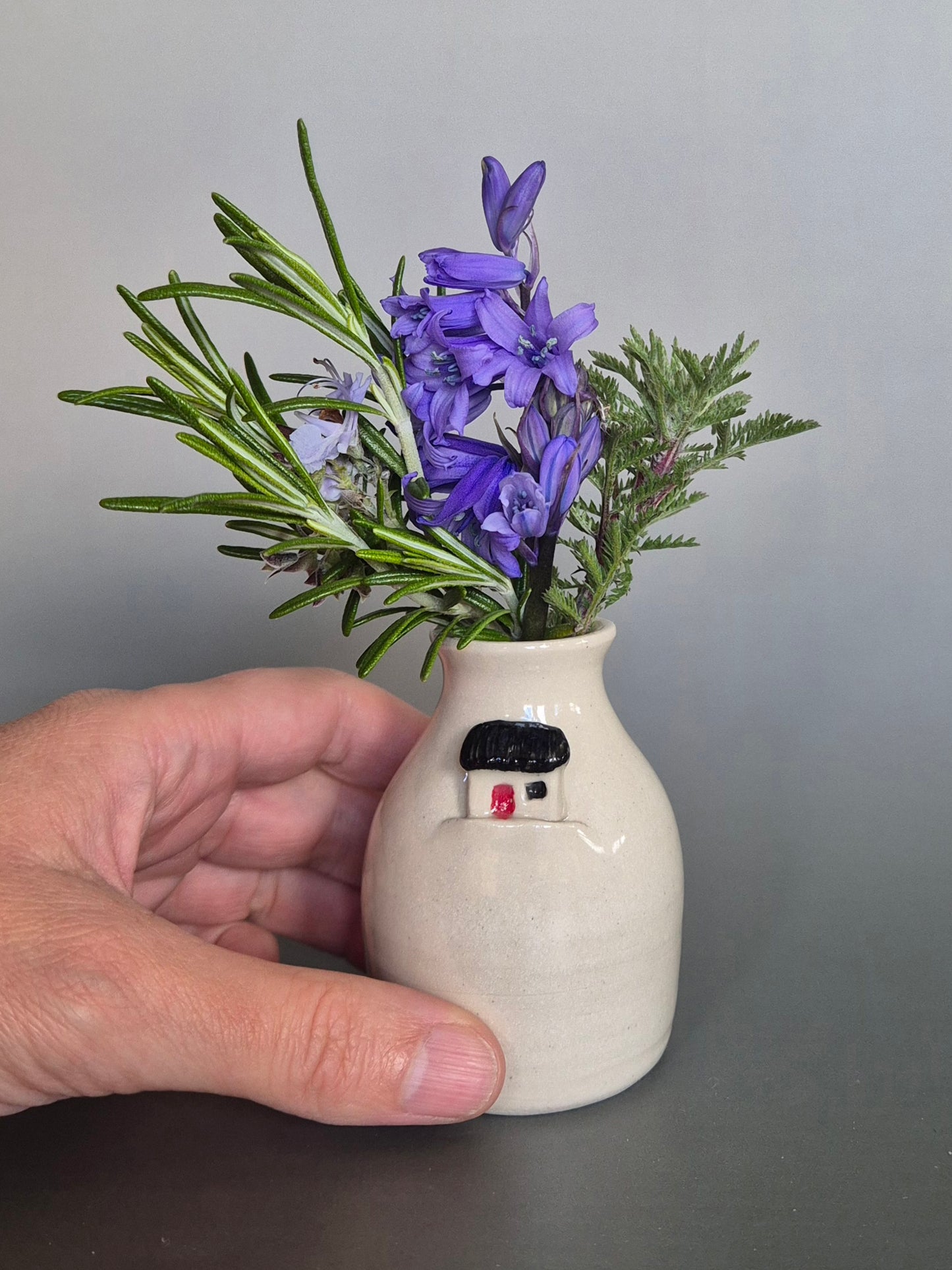 Hand holding a small white ceramic bottle or bud vase decorated with a small house and filled with wildflowers. On a plain background