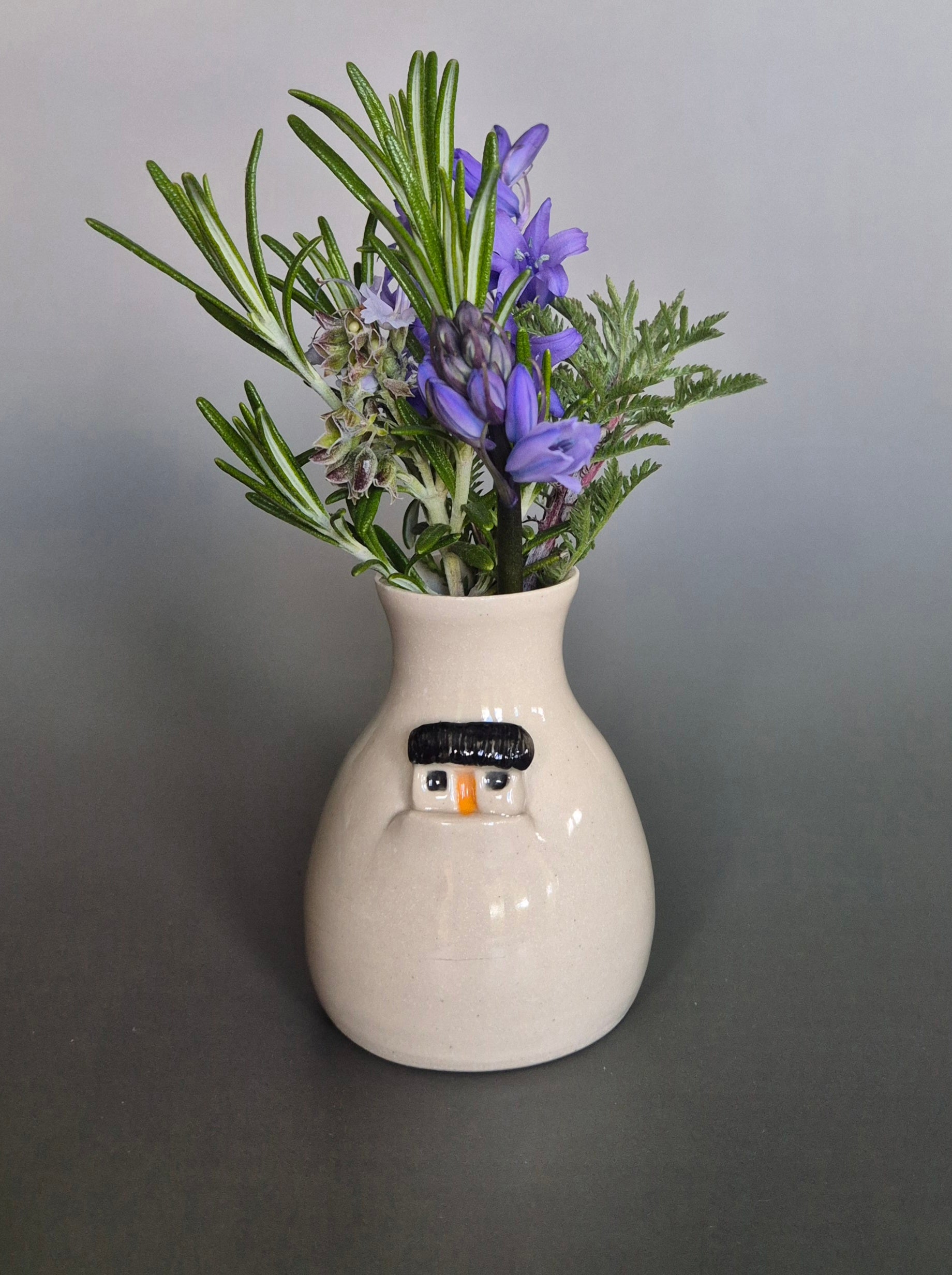 Small white ceramic bottle or bud vase decorated with a small house and filled with wildflowers. On a plain background